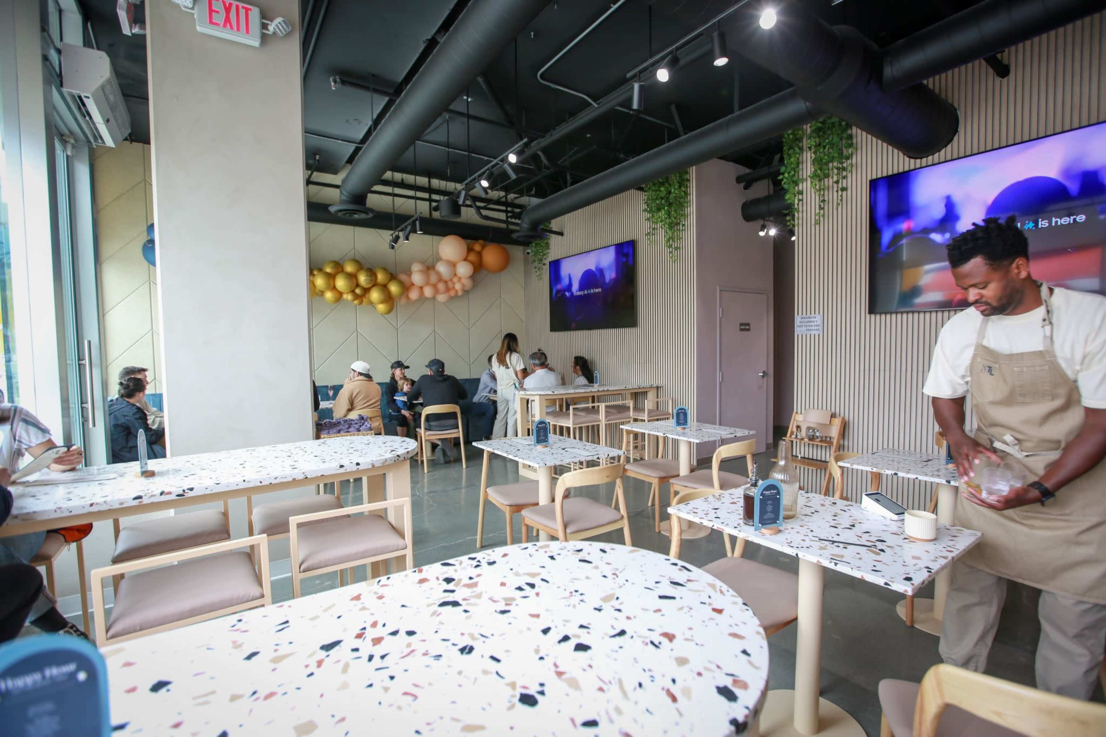 A modern cafe features several tables with colorful terrazzo surfaces, a barista preparing drinks, and customers seated in the background.