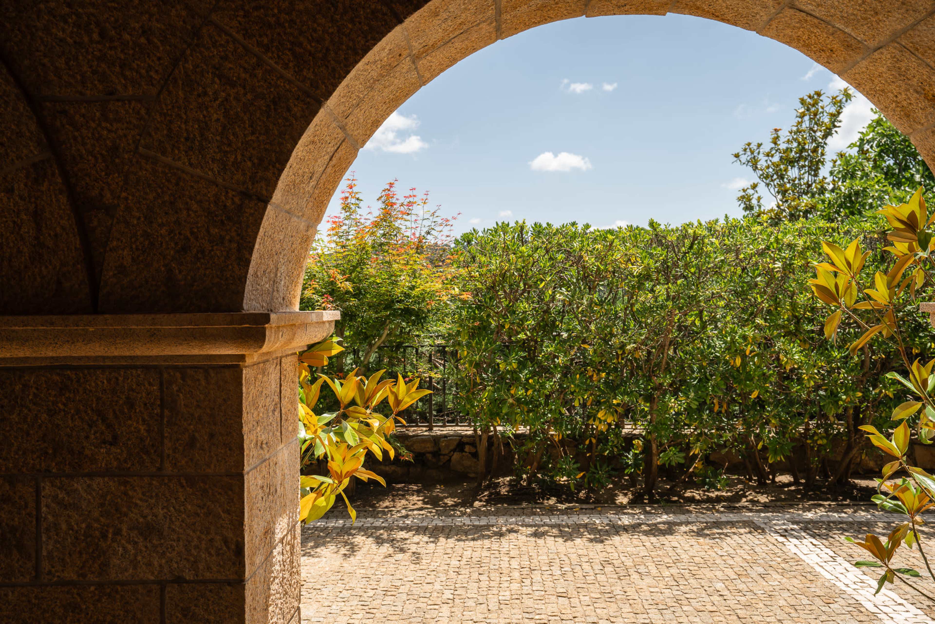 A view through a stone archway, framing a garden with green shrubs and trees under a blue sky.