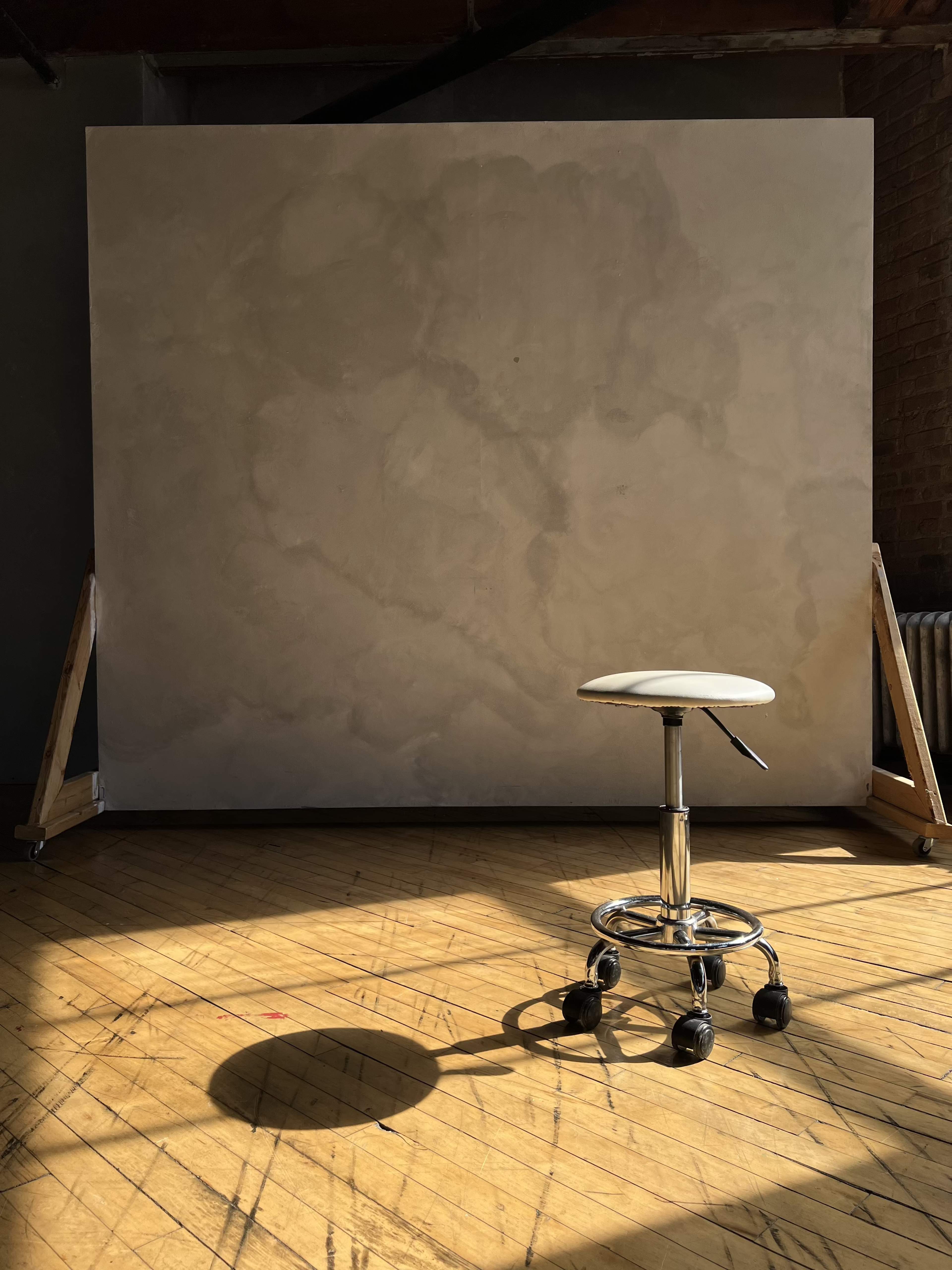 A solitary stool is positioned in front of a large, blank canvas in a well-lit studio space.