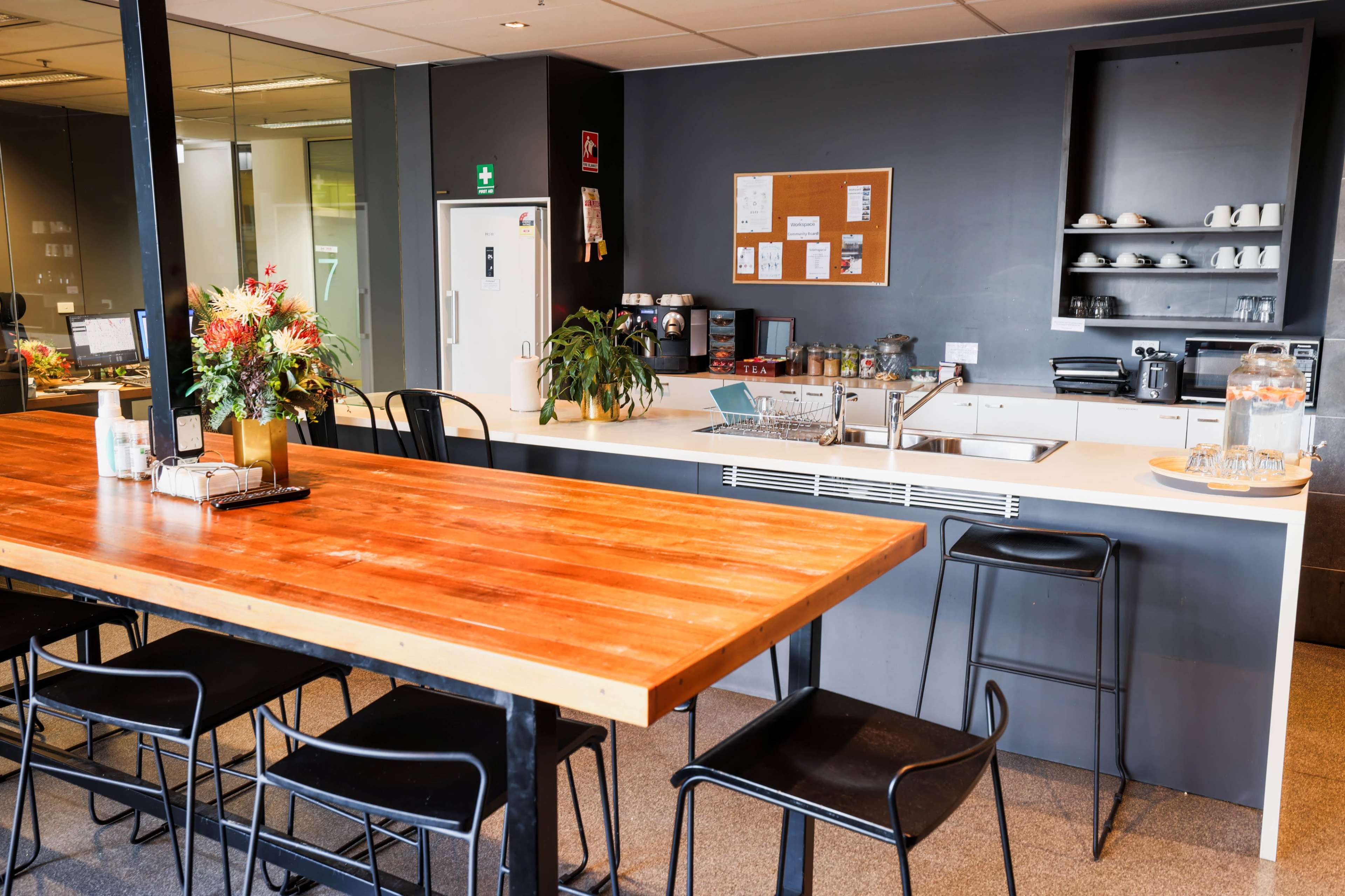 The image shows a modern kitchen area with a large wooden table and black metal chairs, surrounded by plants and kitchen appliances on a counter.