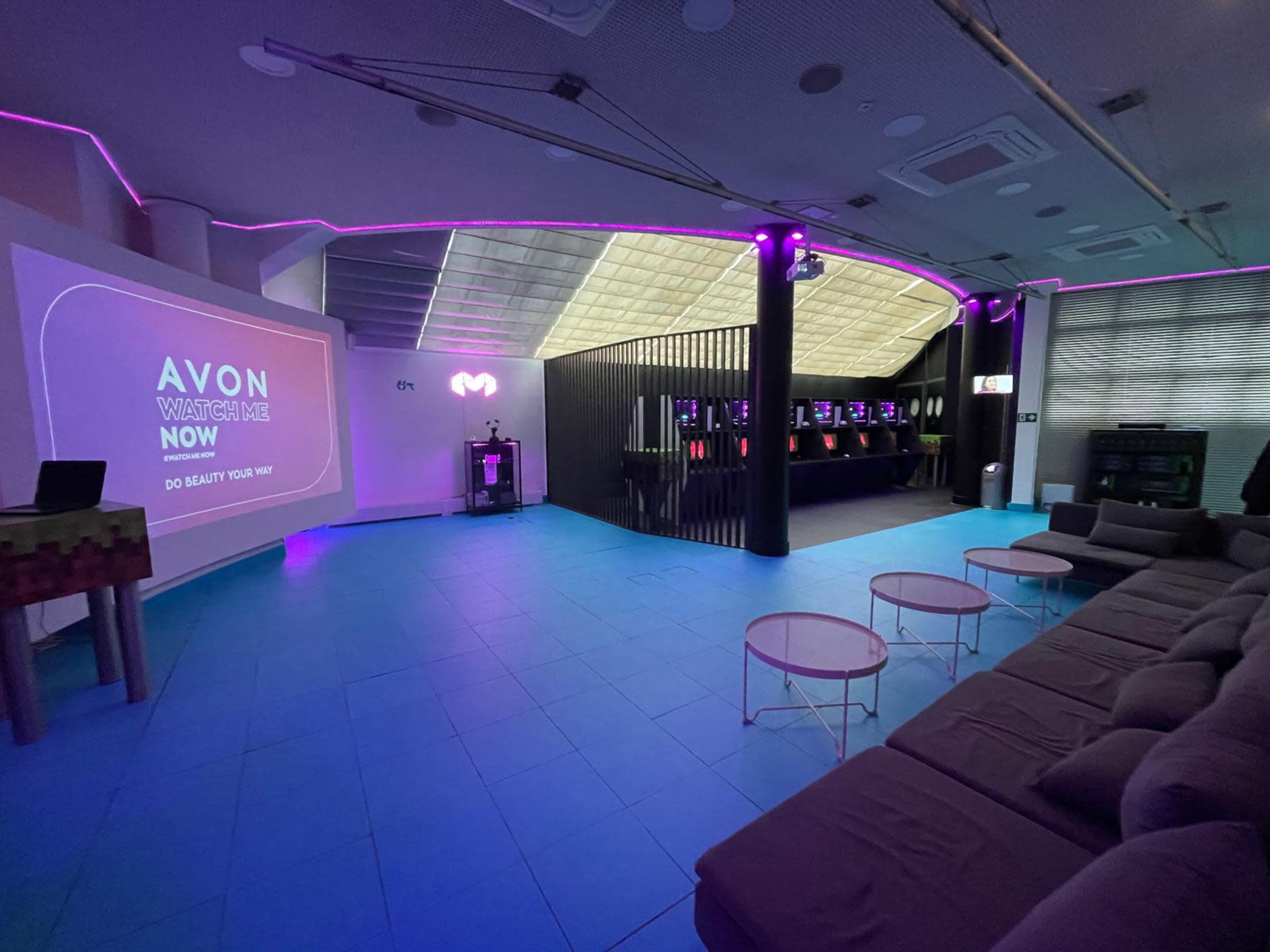 The image shows a modern event space with a large presentation screen on one wall, seating area, and a bar-like setup illuminated with colorful lights.