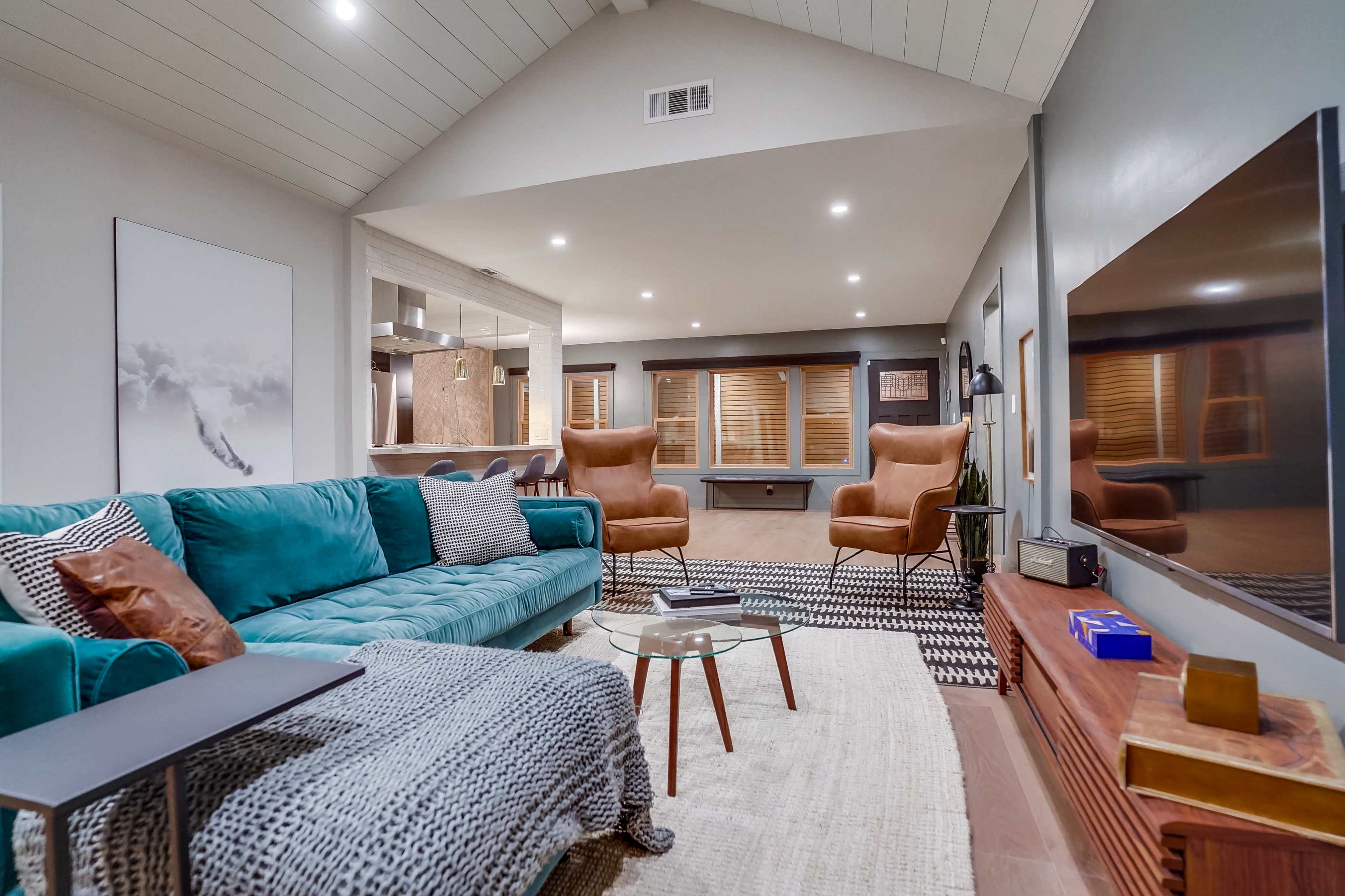 A modern living room featuring a teal sectional sofa, a glass coffee table, and large windows with wooden blinds.