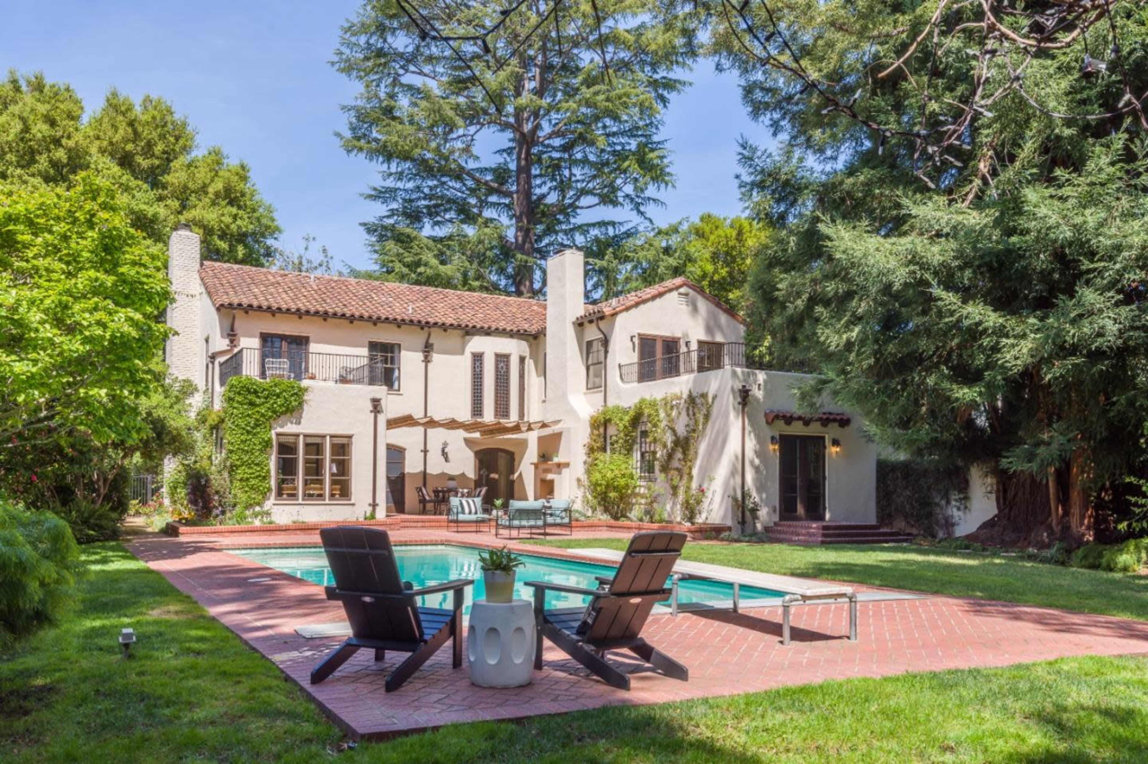 A two-story house with a tiled roof and a swimming pool surrounded by lounge chairs in a landscaped backyard.