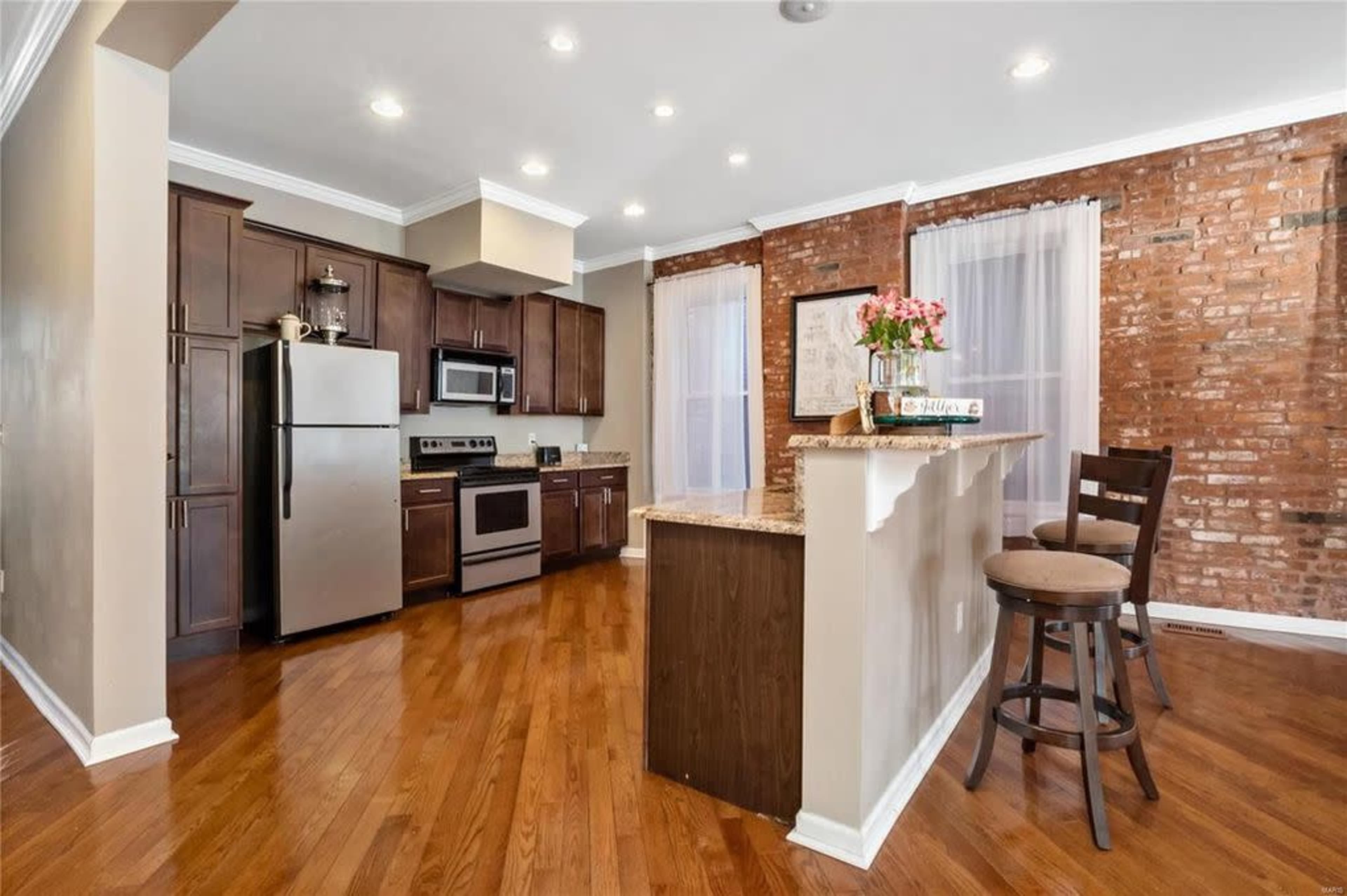 A modern kitchen with stainless steel appliances, dark wood cabinets, a granite countertop, and hardwood flooring, illuminated by recessed lighting.