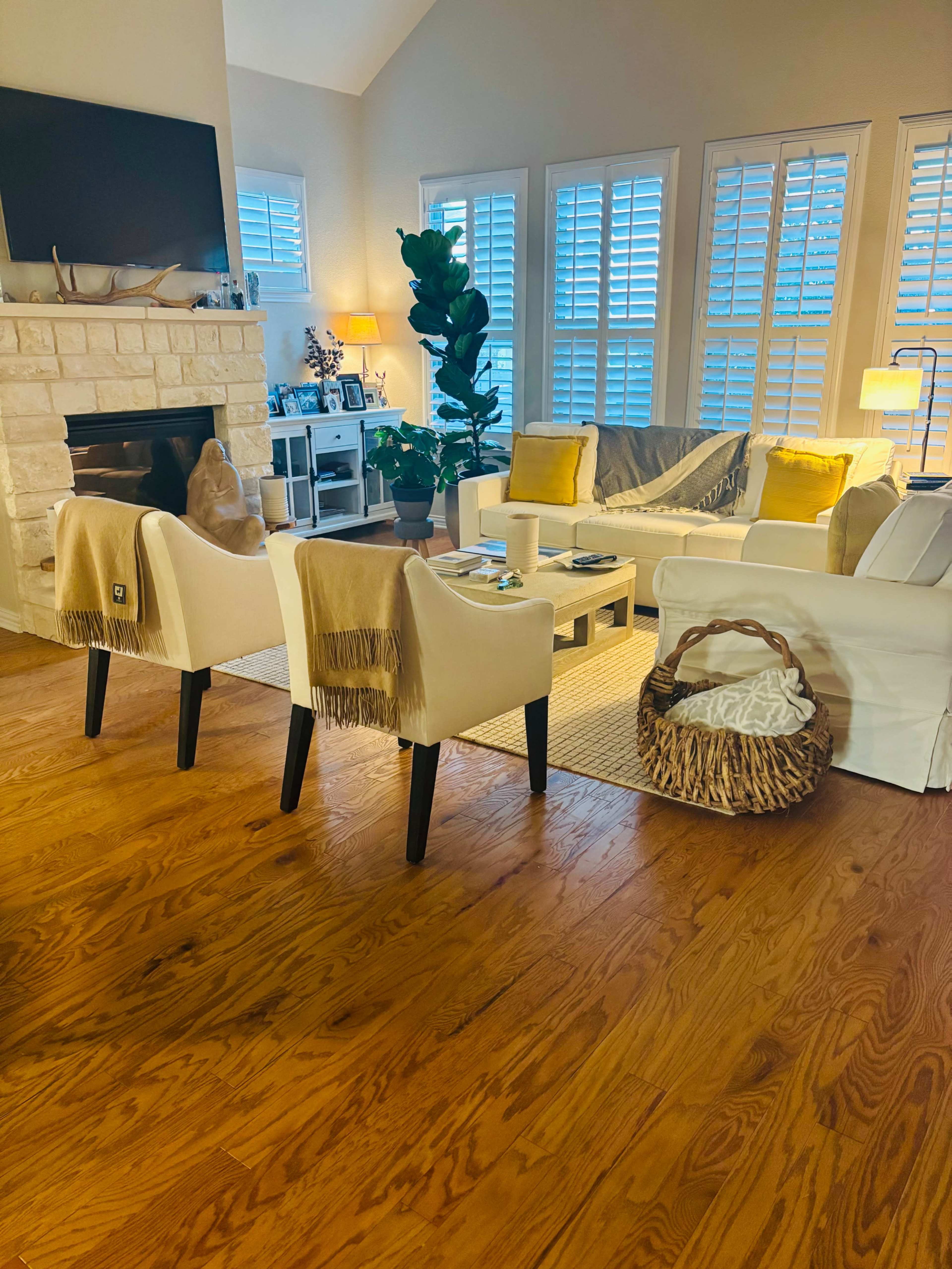 The image shows a cozy living room with a stone fireplace, light-colored furniture, and large windows covered with shutters.