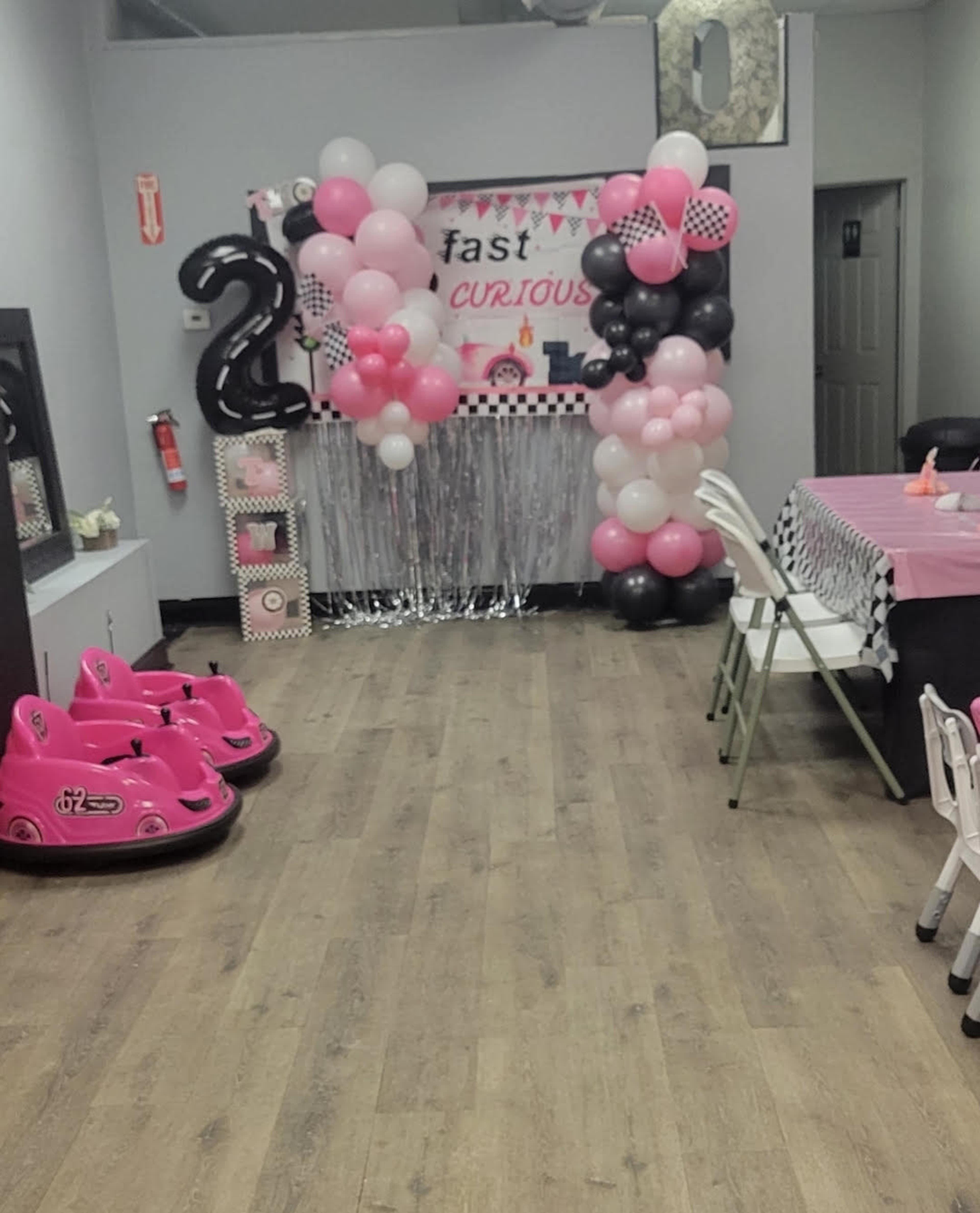 The image shows a decorated room for a birthday party featuring balloon arches in pink and black, a table with a pink tablecloth, and two pink toy cars positioned at the front.