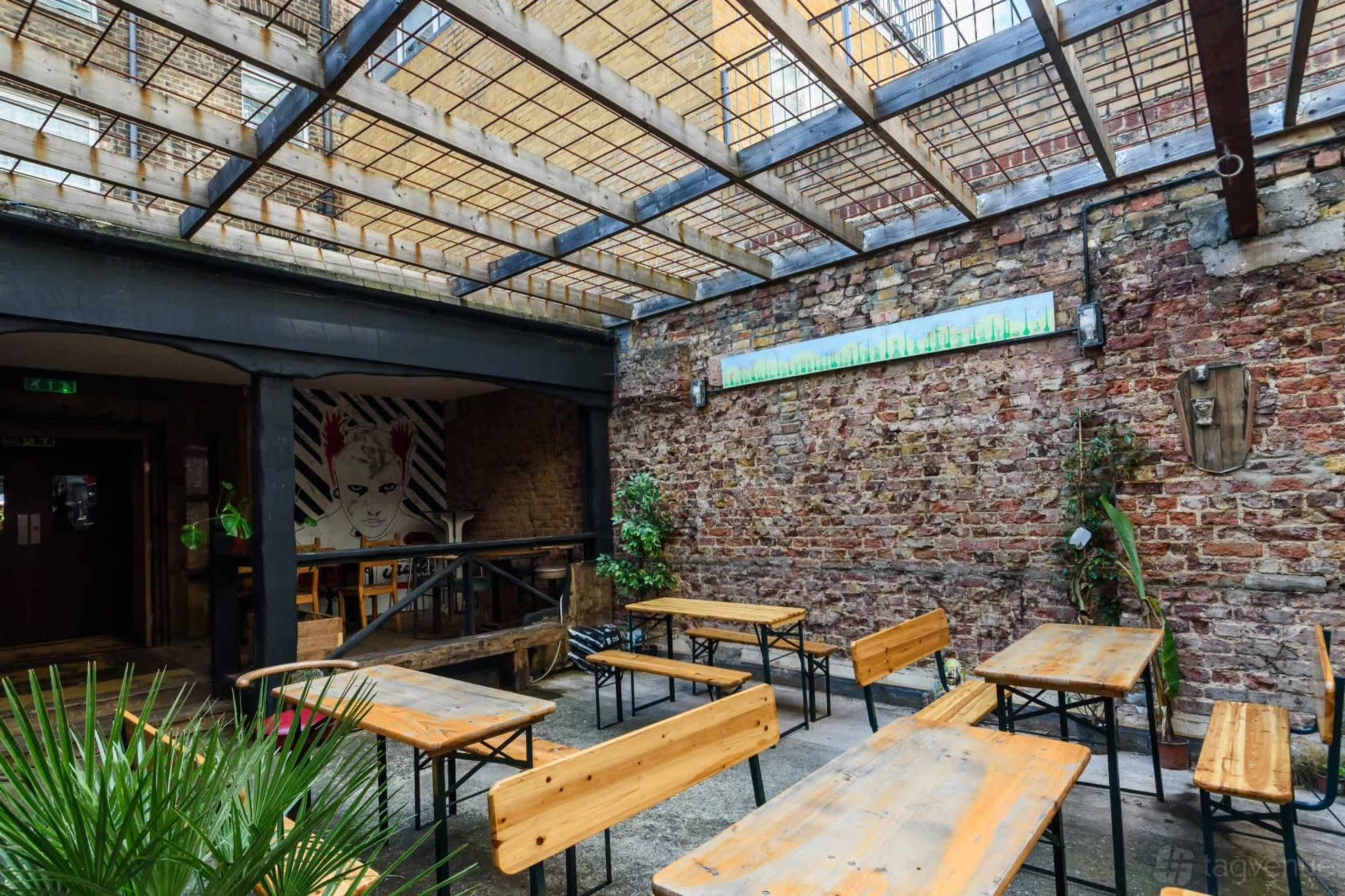 The image shows an indoor space with wooden benches and tables under a glass roof, featuring exposed brick walls and greenery.