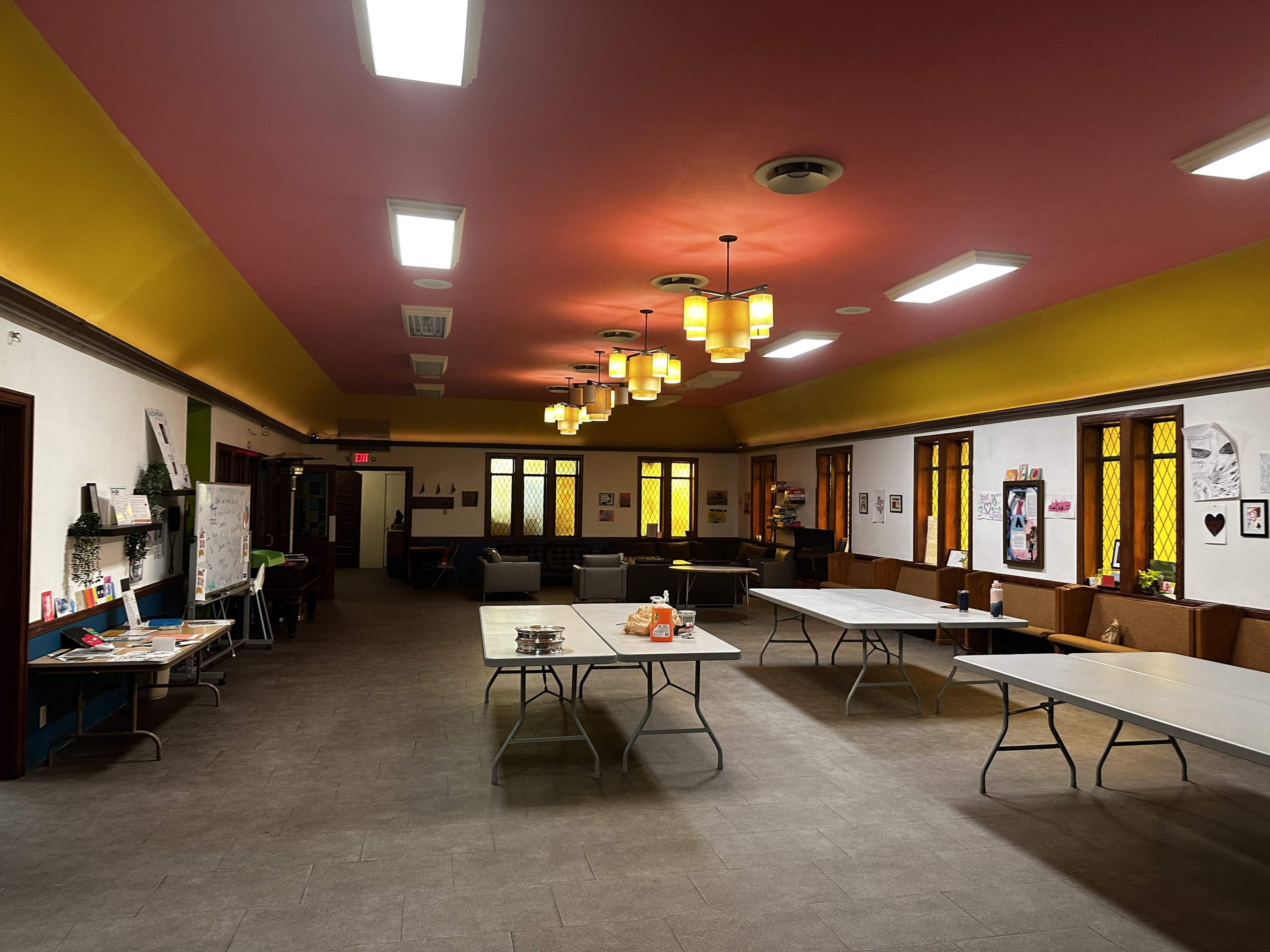 A spacious indoor room with tables arranged in the center, chairs along the walls, and decorative lighting above.