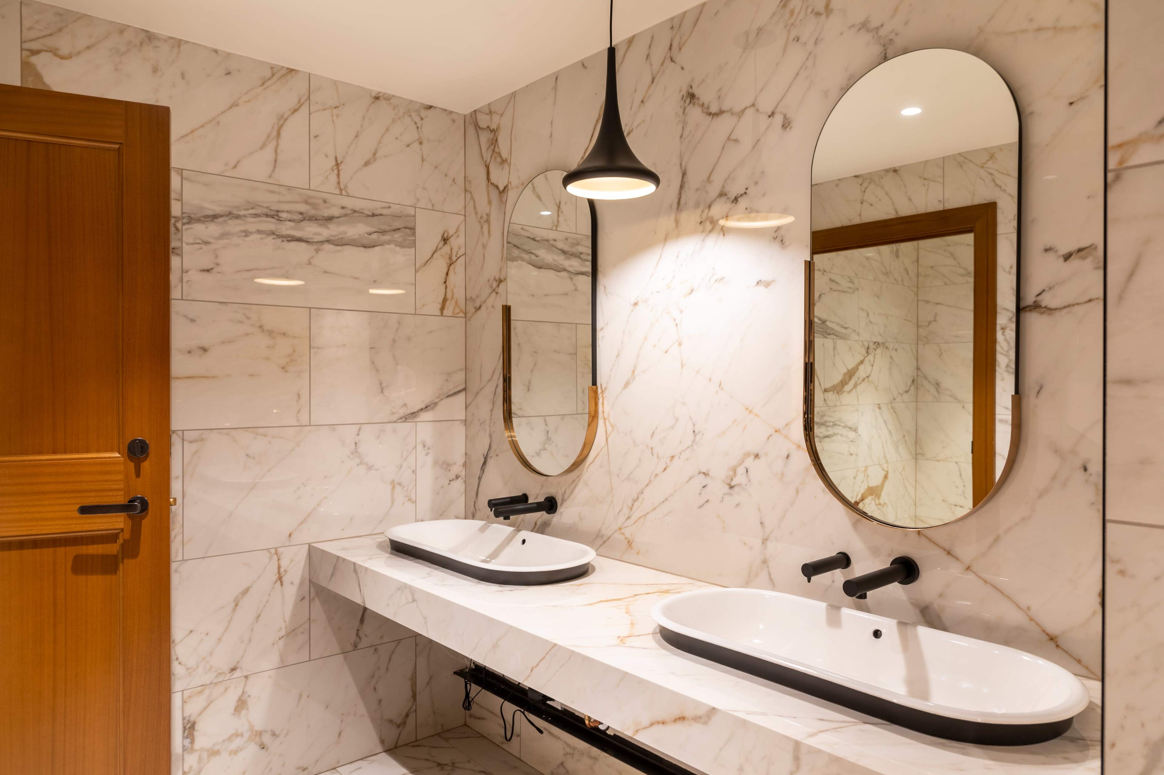 A modern bathroom with marble walls, two oval mirrors above a double sink, and a wooden door.