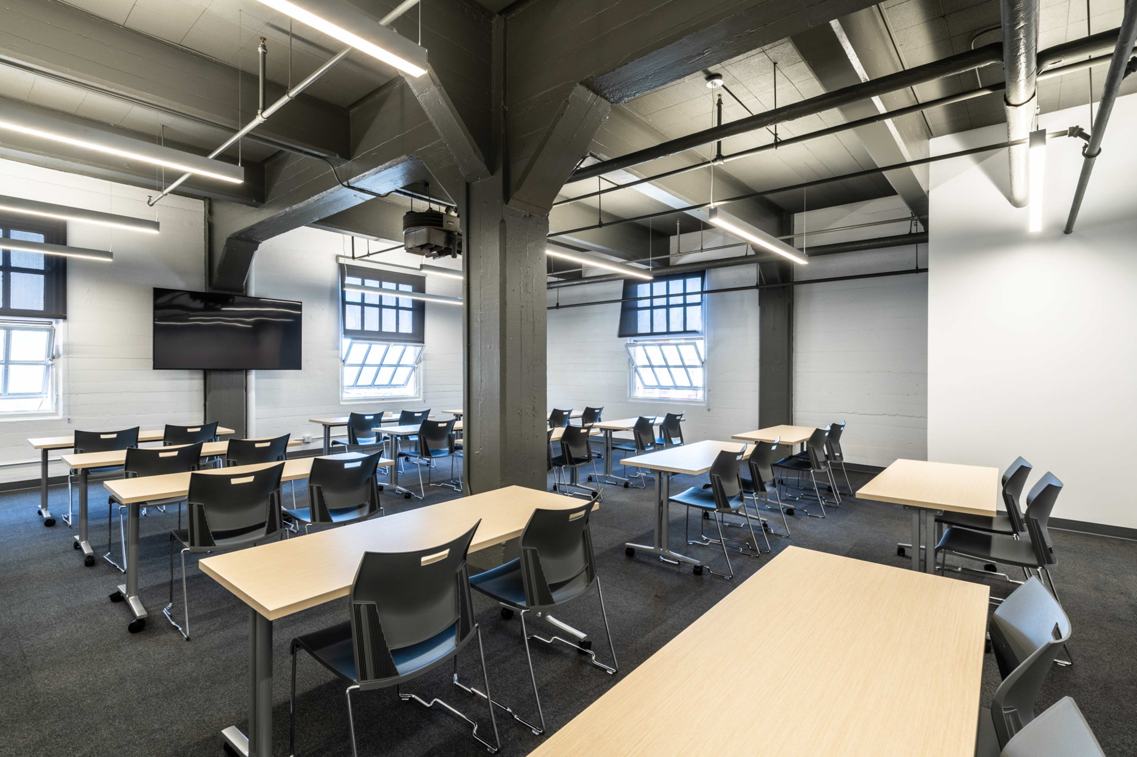 A spacious classroom with several rows of tables and chairs, a wall-mounted screen, and large windows allowing natural light.