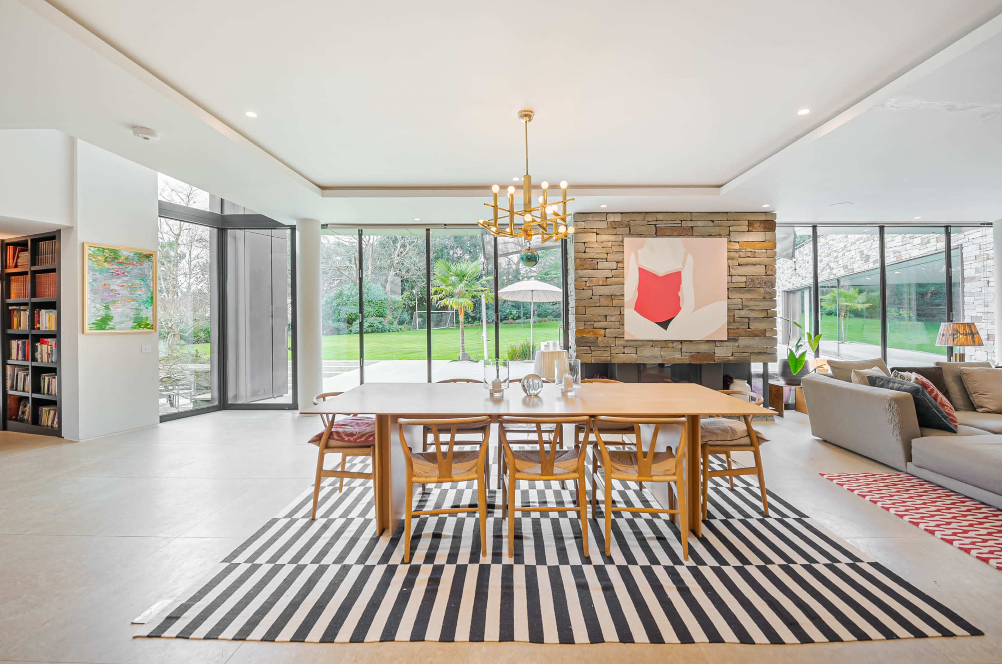 A modern dining area features a wooden table surrounded by chairs, a stone accent wall with artwork, and large windows overlooking a garden.