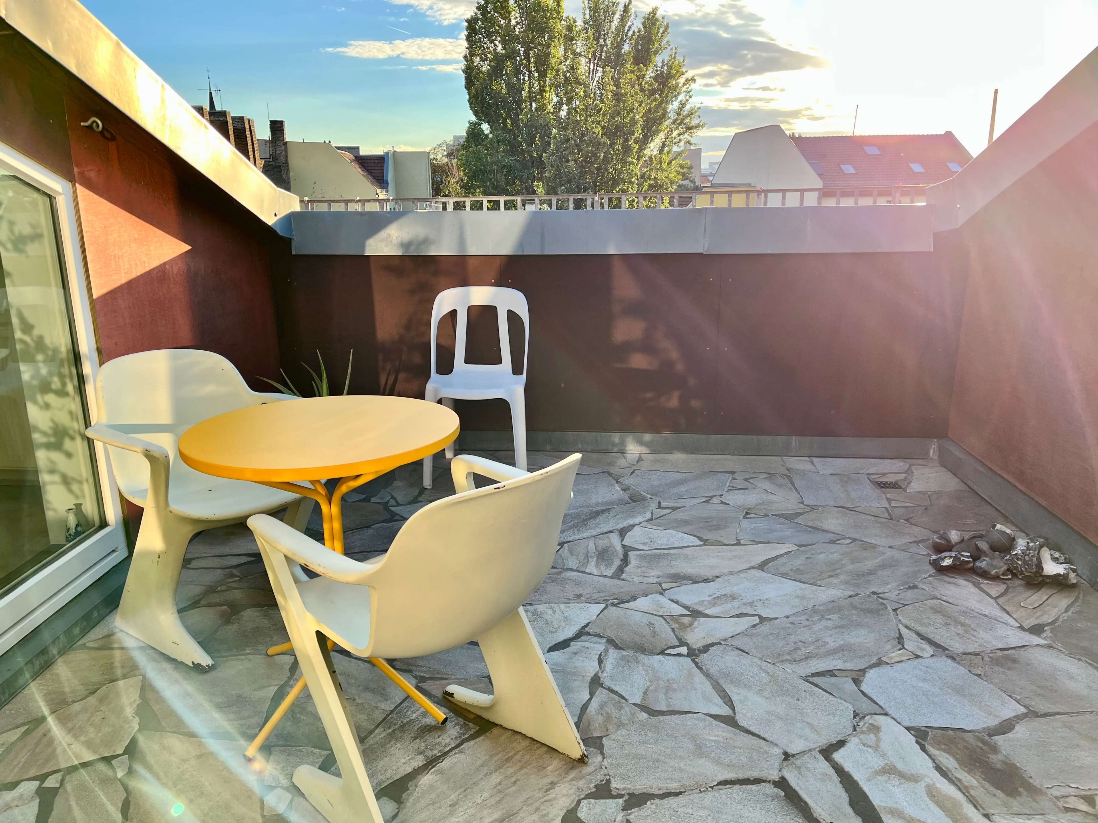 A rooftop terrace with a round yellow table and three white chairs surrounded by a stone floor and greenery.