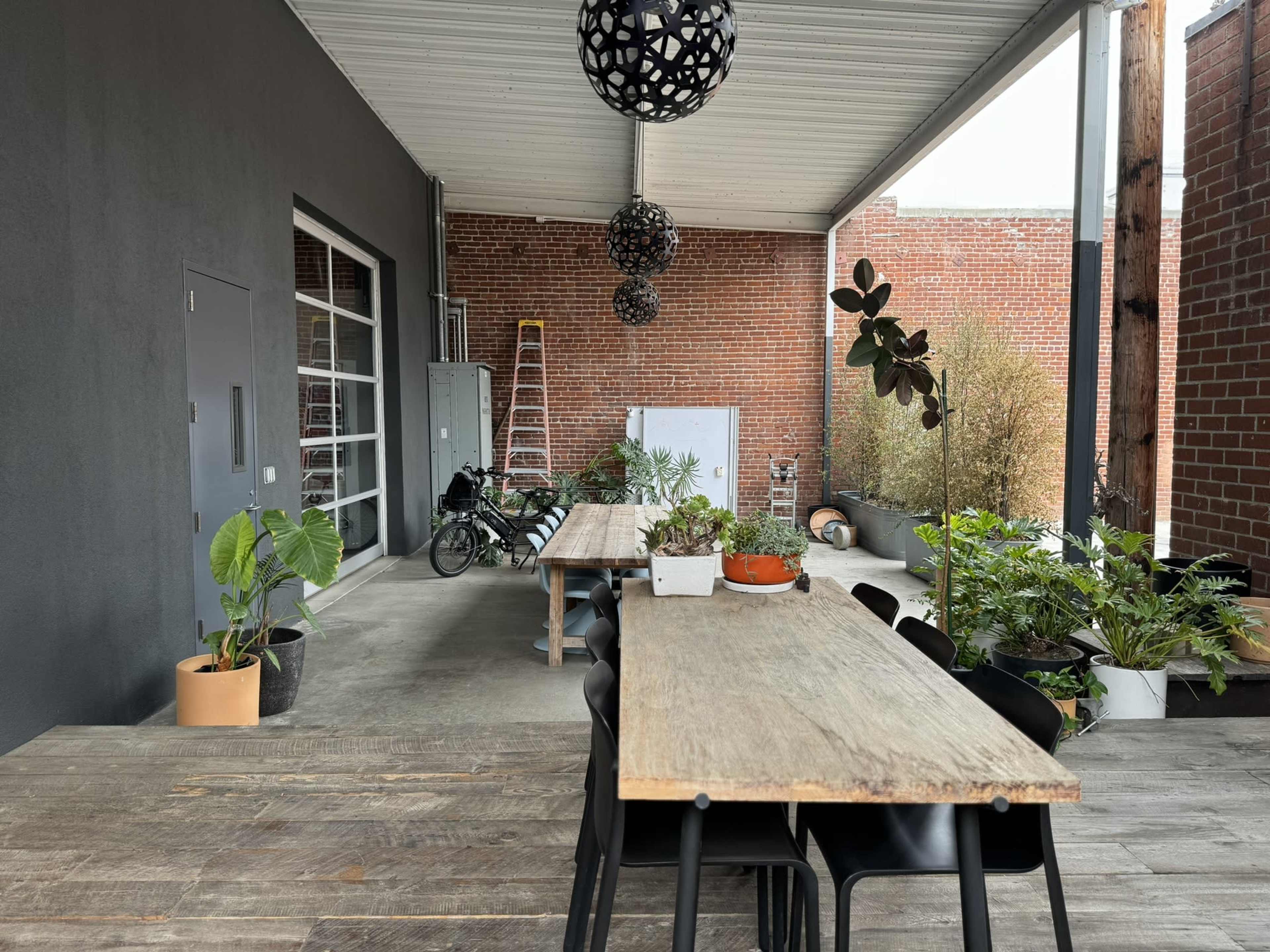 The image shows a covered outdoor area with wooden tables, black chairs, and various potted plants against a brick wall.