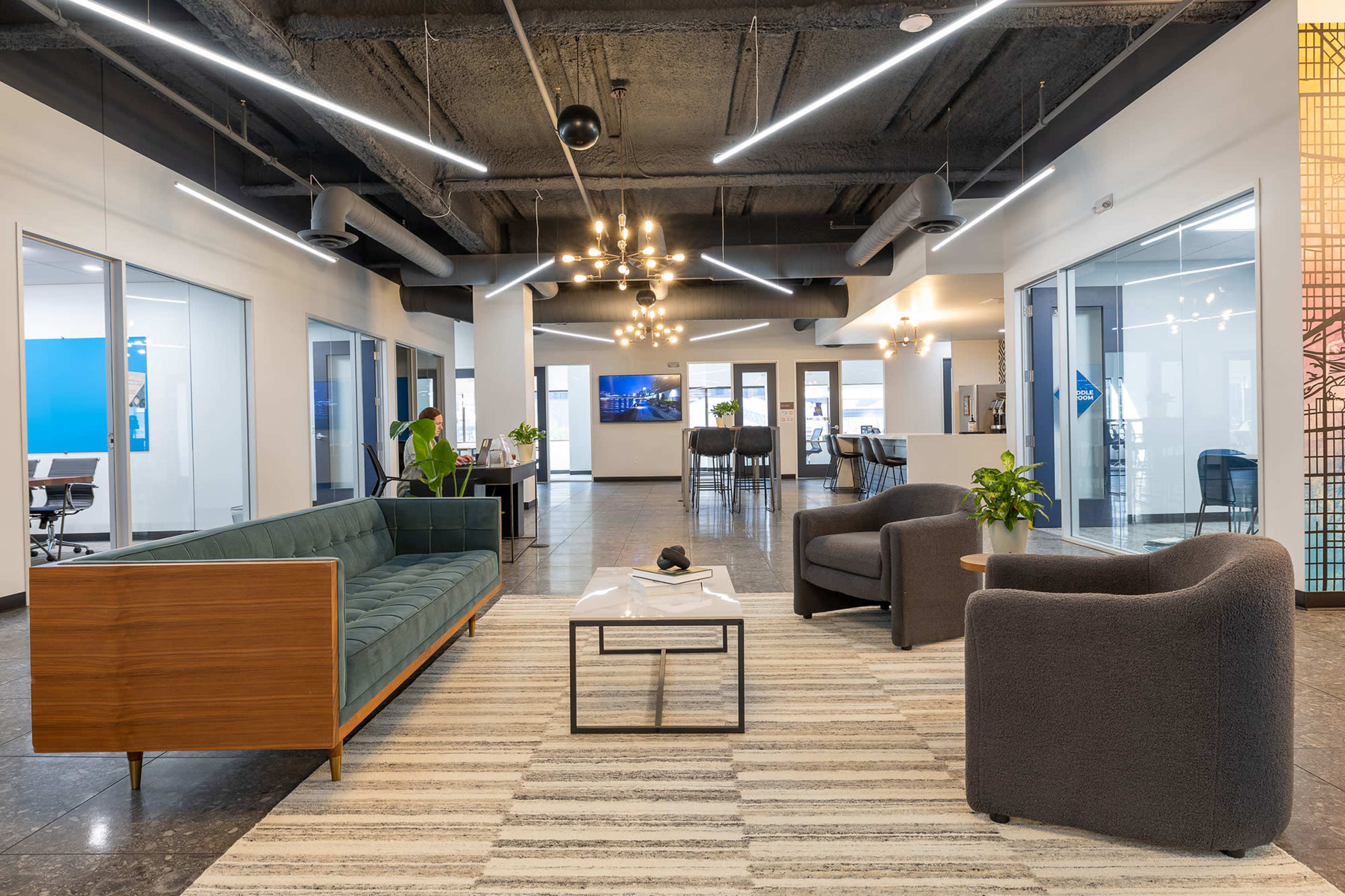 The image shows a modern office lounge area furnished with a green couch, two gray armchairs, a coffee table, and decorative plants, all under a contrasting ceiling with exposed beams and modern lighting fixtures.