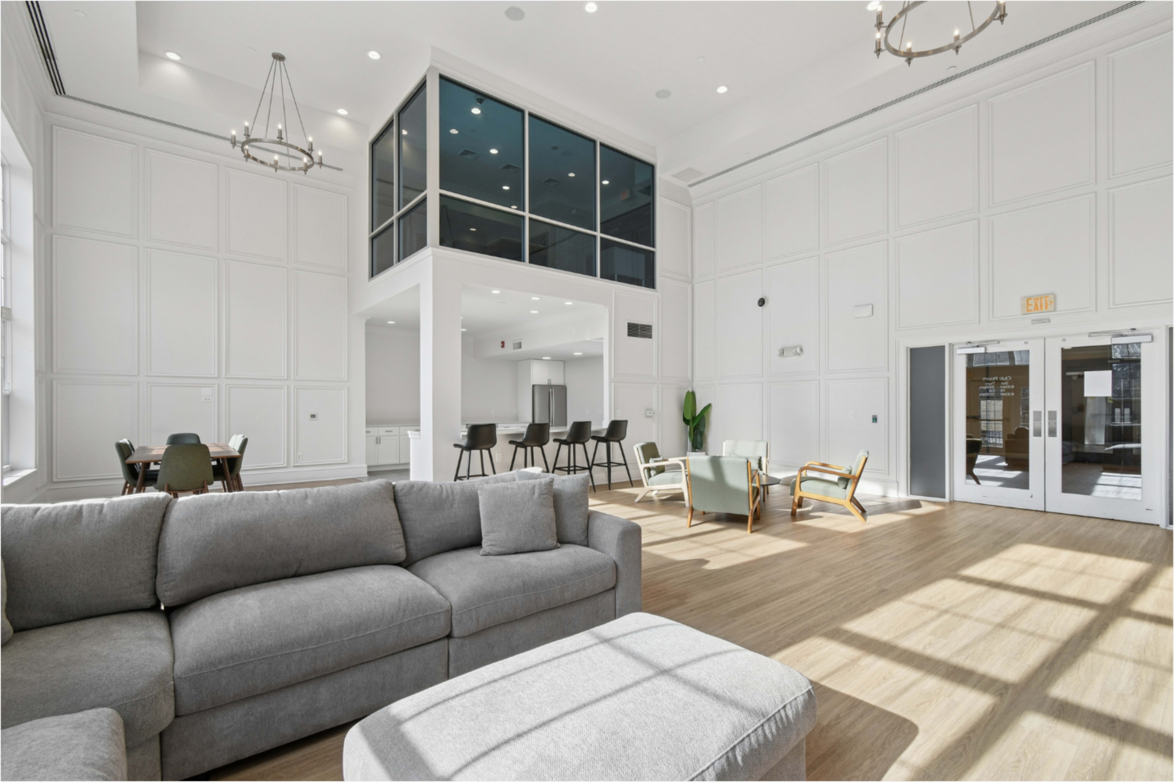The image shows a modern, well-lit interior space with white walls, large windows, and a combination of seating areas that includes a gray sectional sofa and several chairs.