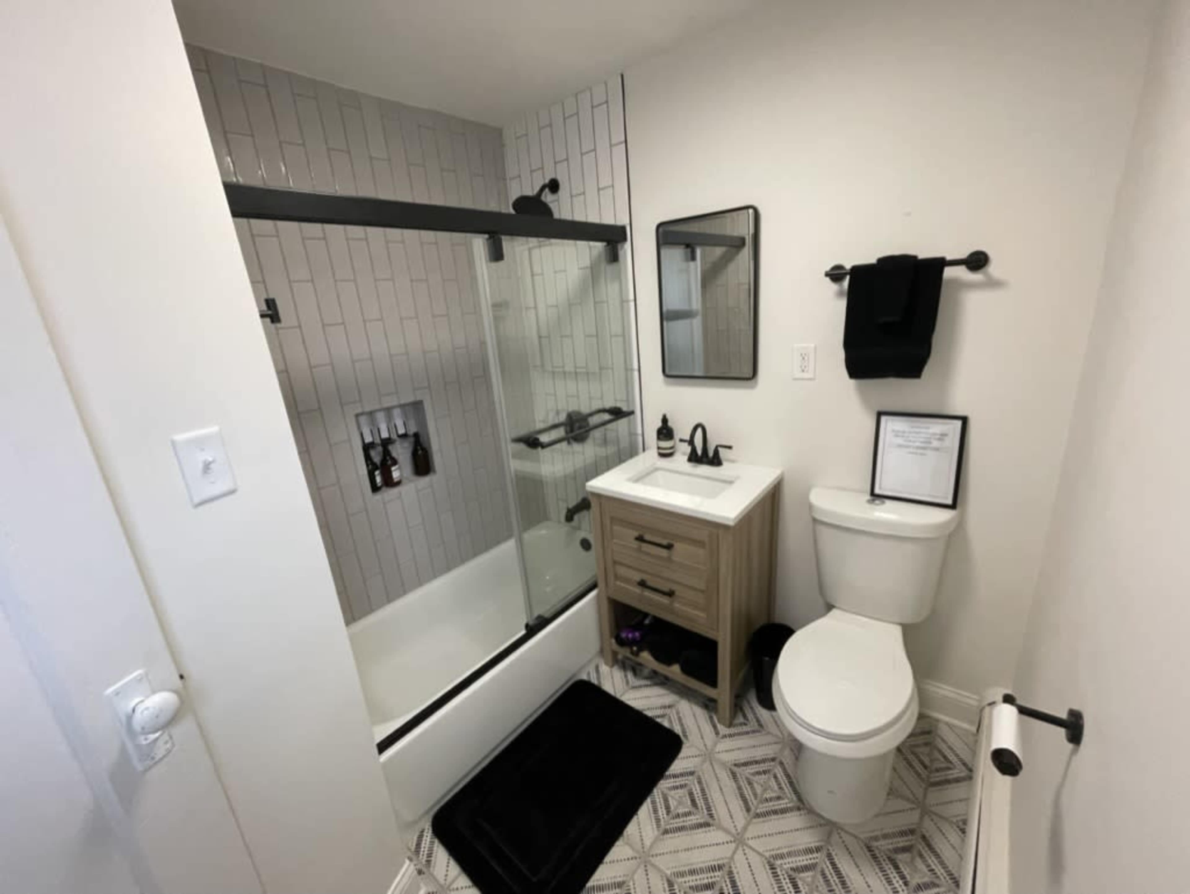 The image shows a modern bathroom featuring a glass shower enclosure, a wooden vanity with a sink, a toilet, and geometric-patterned tile flooring.