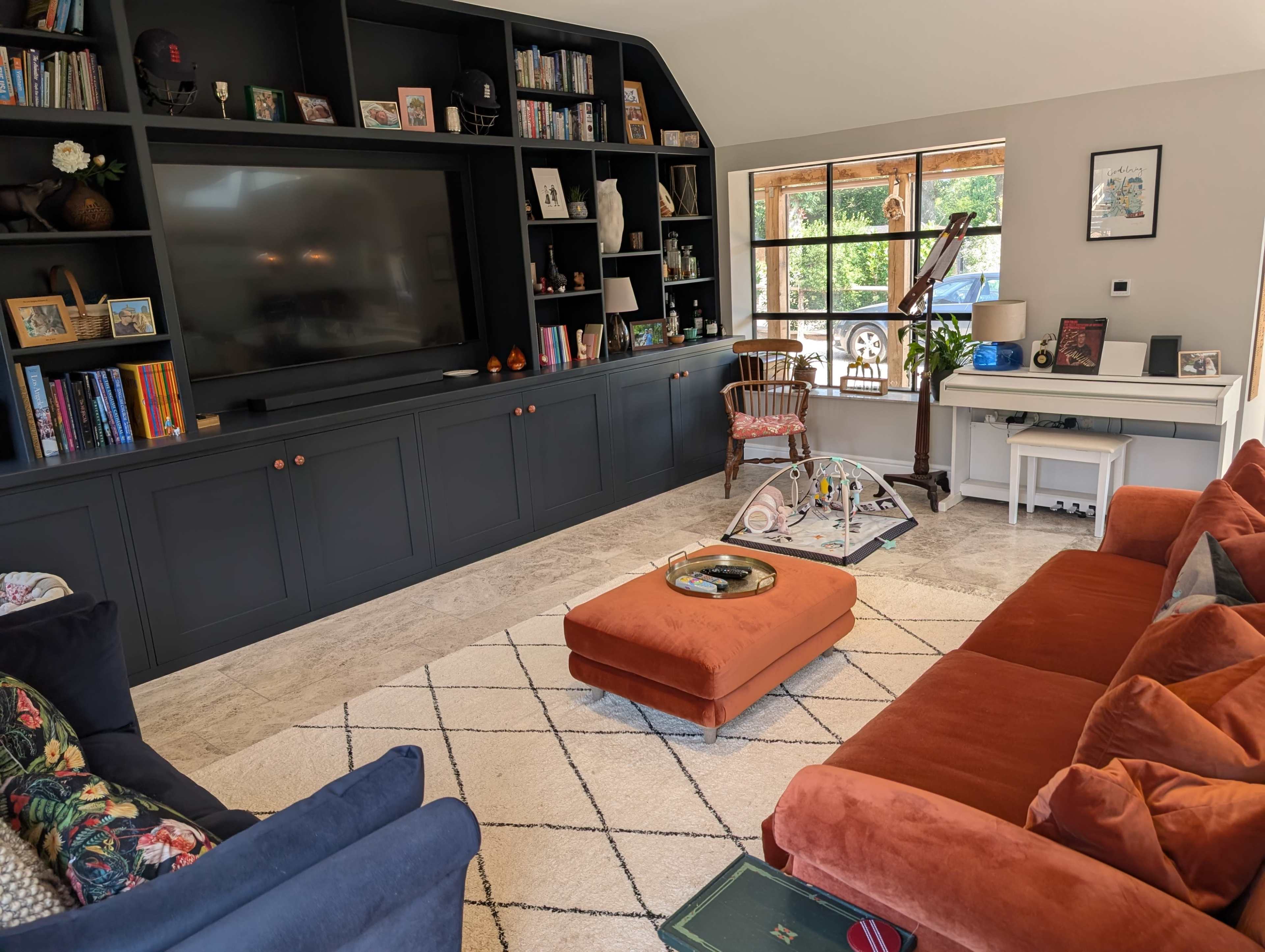 The image shows a cozy living room with a large television, a dark bookshelf filled with books and decor, an orange sofa, a patterned rug, and a desk near a window.