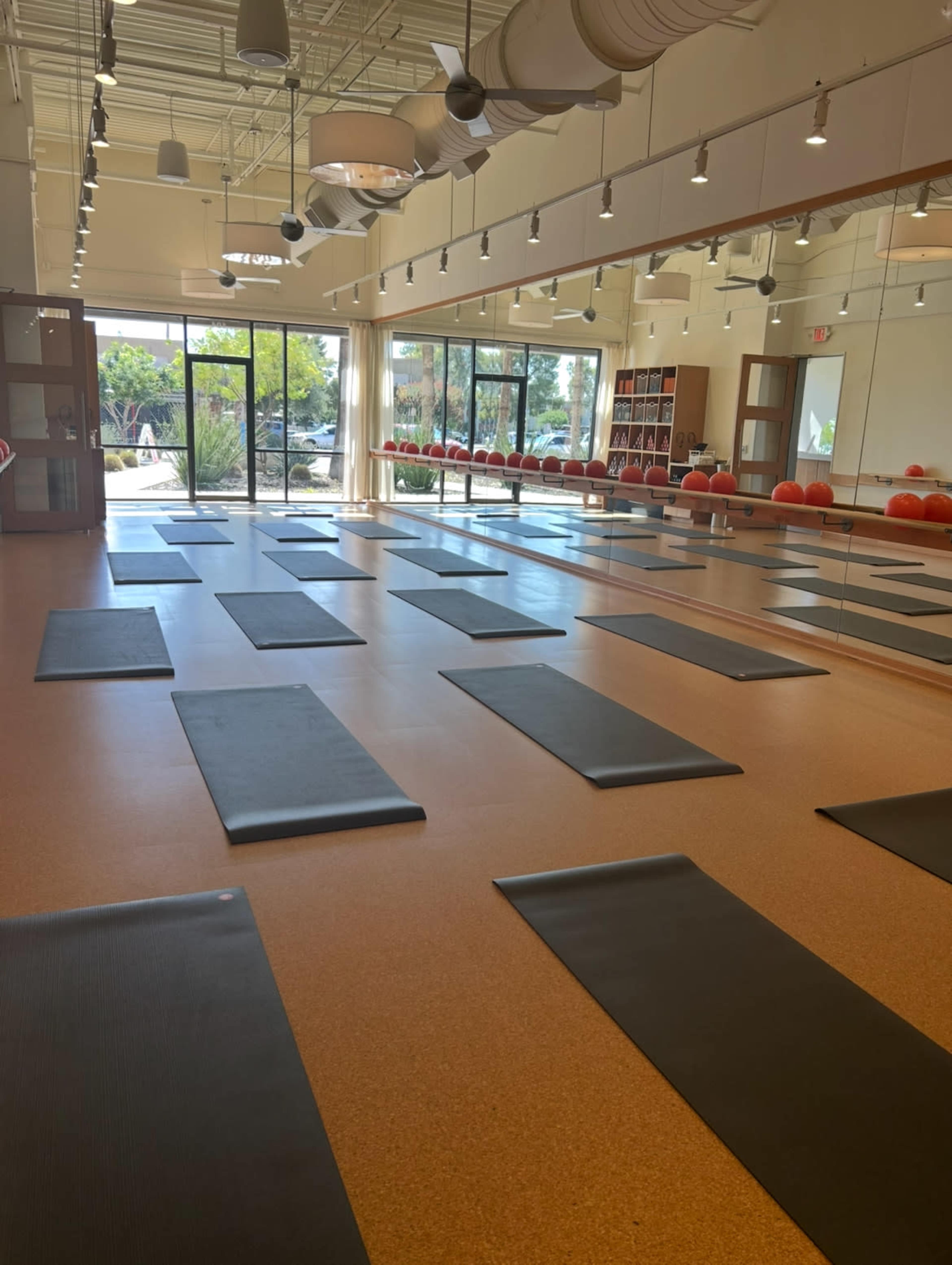 The image shows an empty yoga studio with several black mats arranged on a cork floor, large windows letting in natural light, and storage shelves in the background.