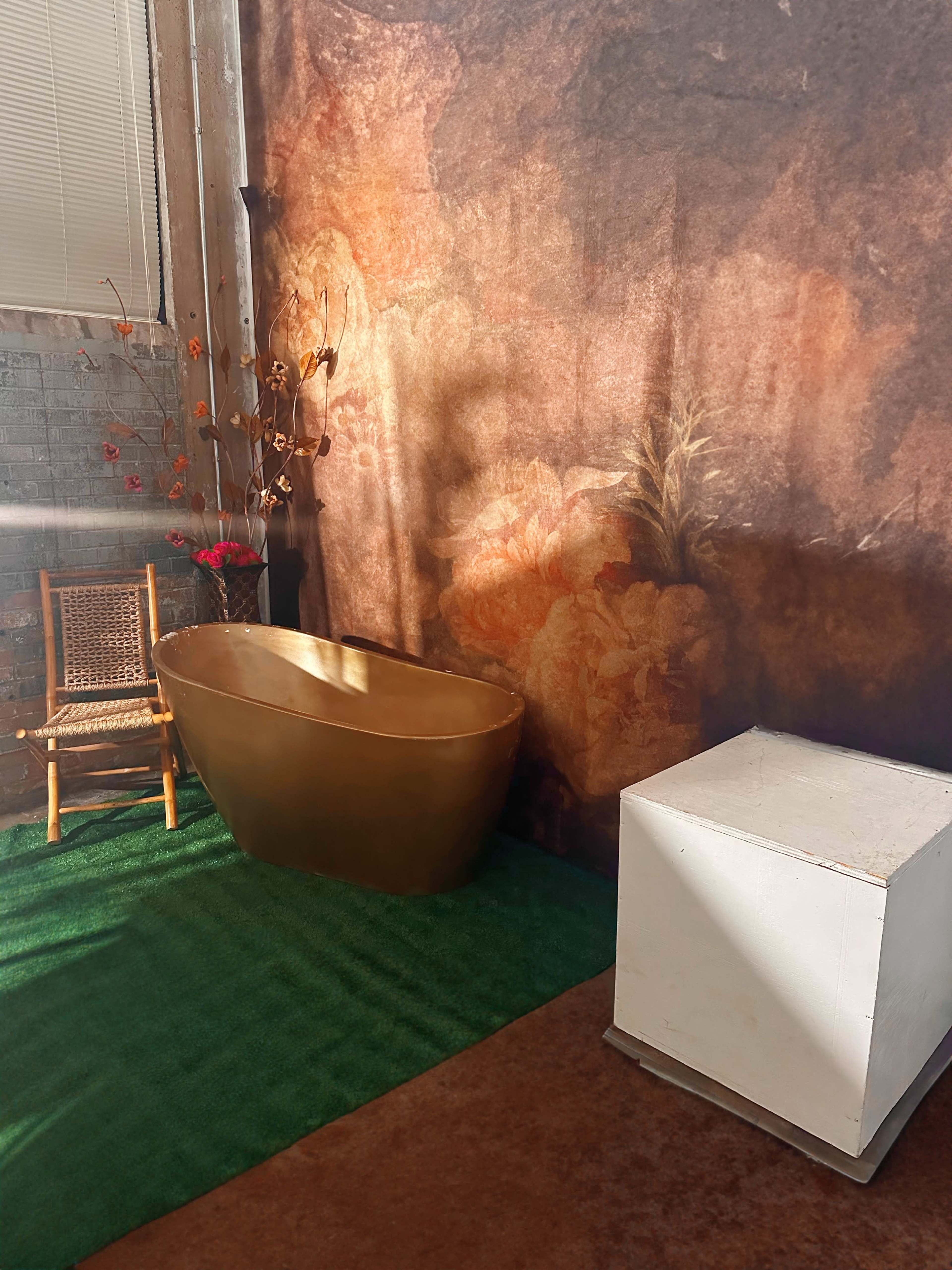 The image shows a small bathroom setup with a golden tub, a white cube table, and a bamboo chair against a floral wall backdrop.