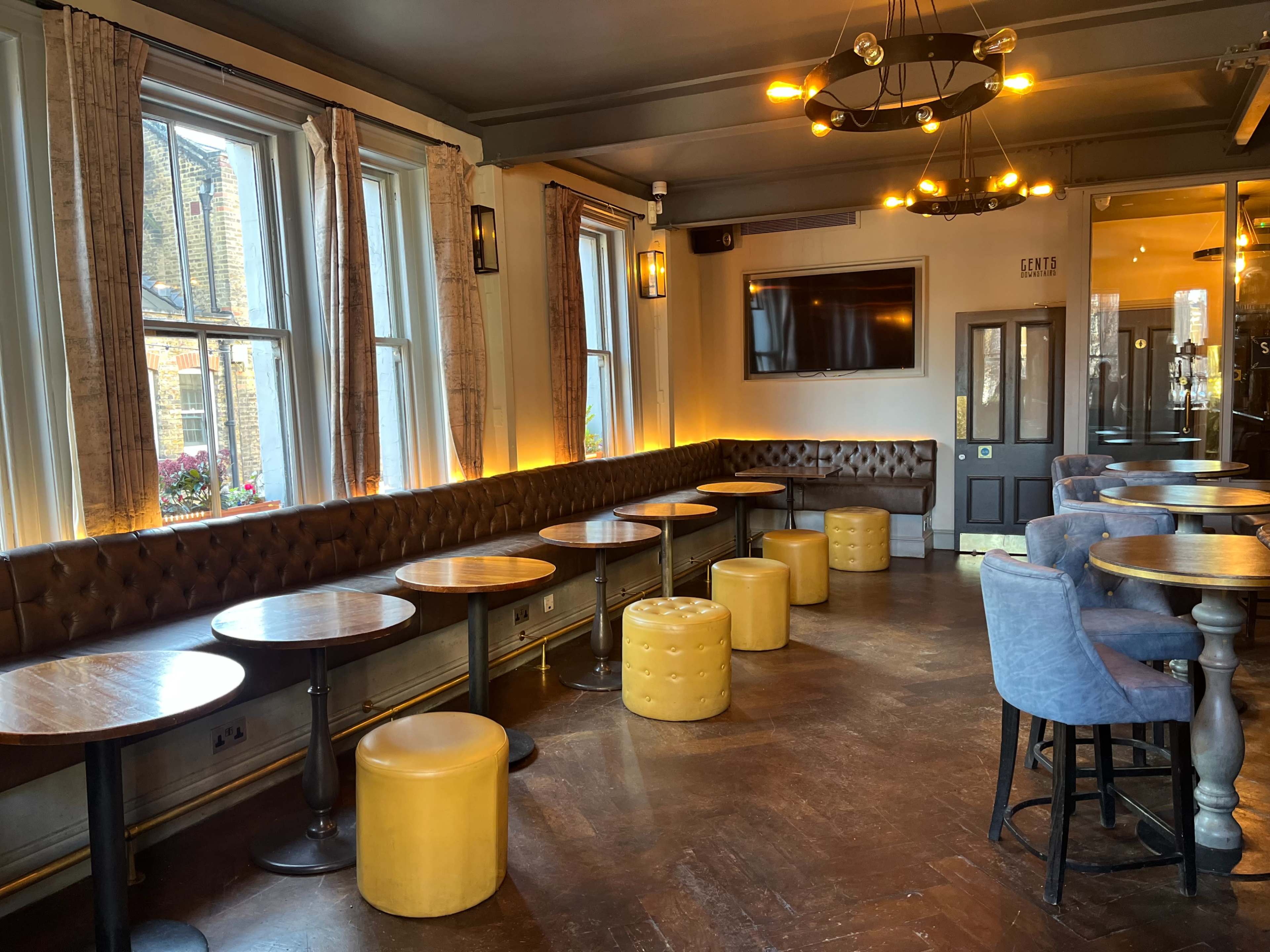 The interior of a pub features a row of tables and yellow upholstered stools arranged along a wall, with large windows allowing natural light to illuminate the space.