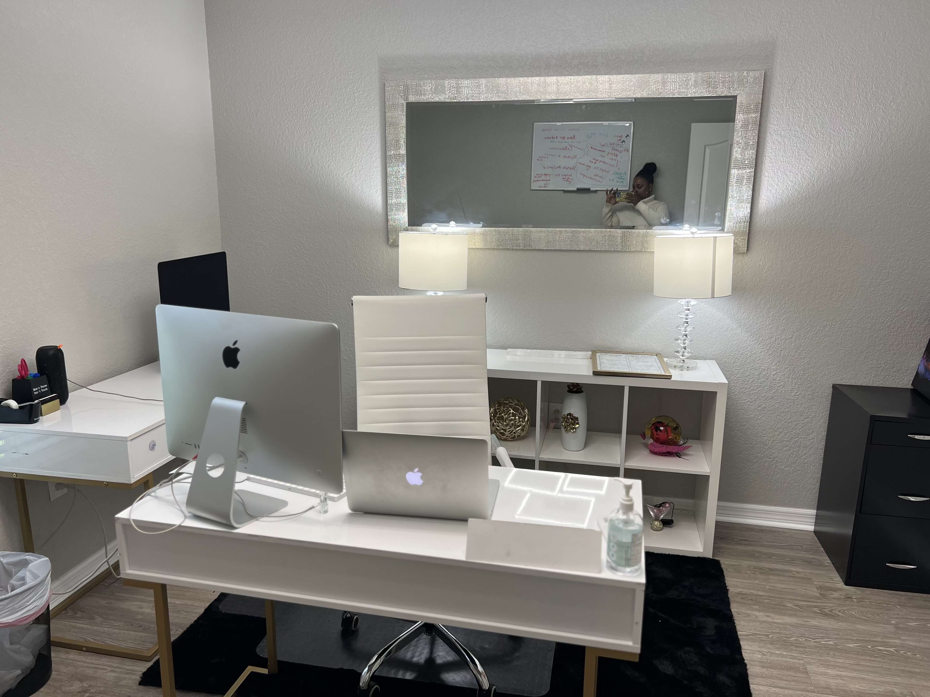 A modern office space features a desk with two Apple computers, a chair, a lamp, and a whiteboard visible in the mirror reflection.