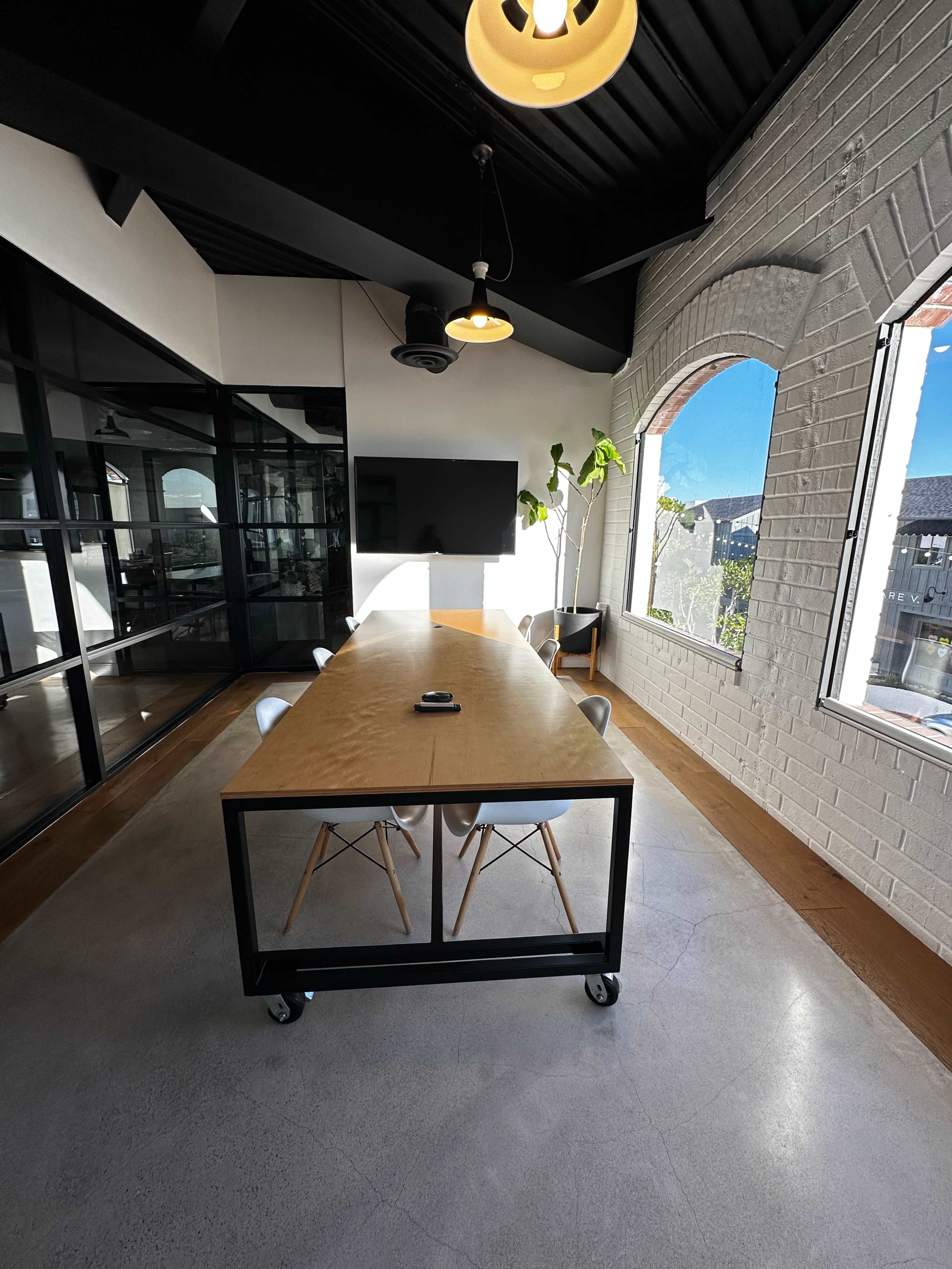 A modern conference room features a long wooden table with rolling chairs, large windows, and a wall-mounted screen.