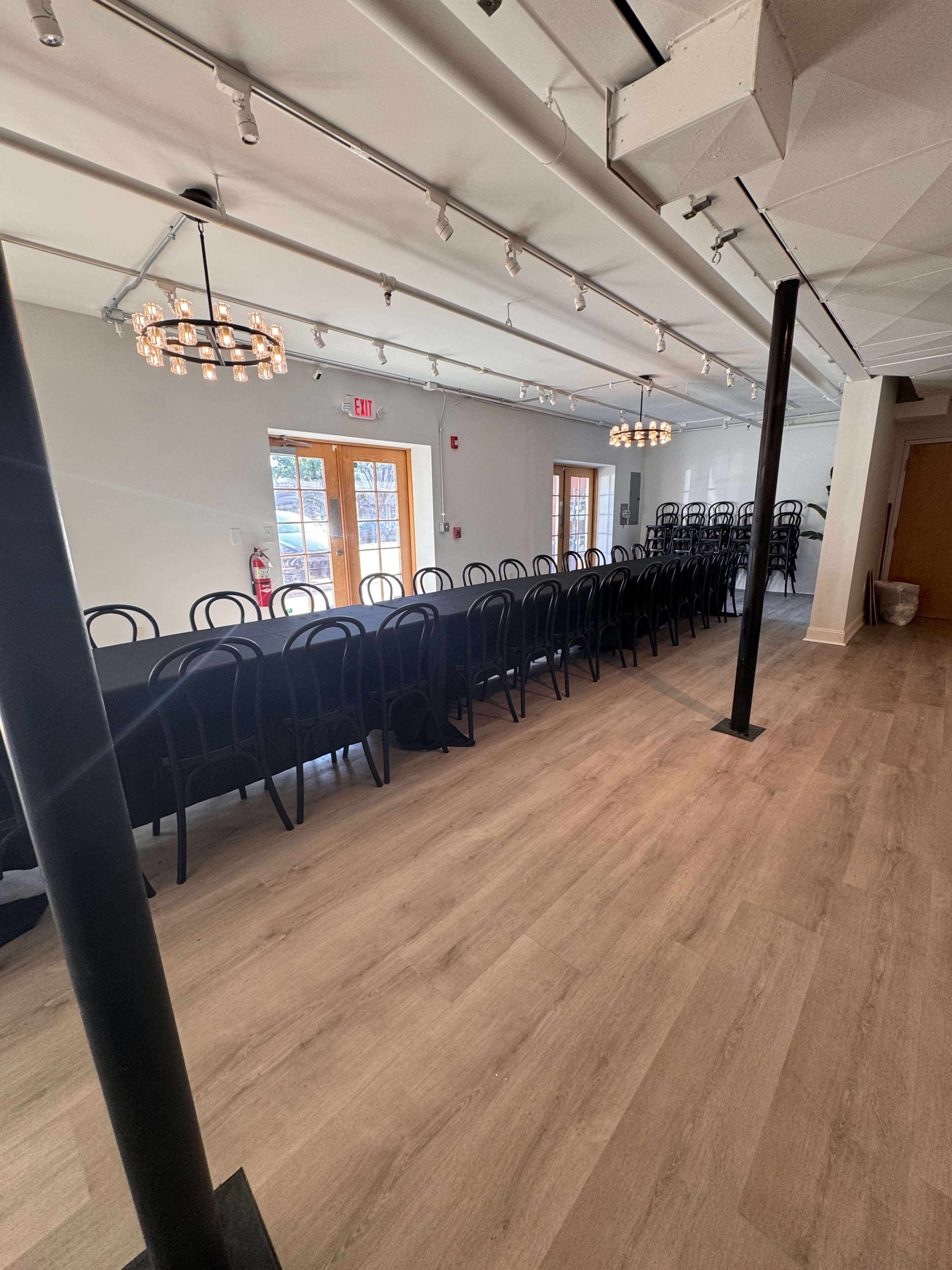 The image shows a spacious event room with a long table set for dining, surrounded by black chairs, and large windows letting in natural light.