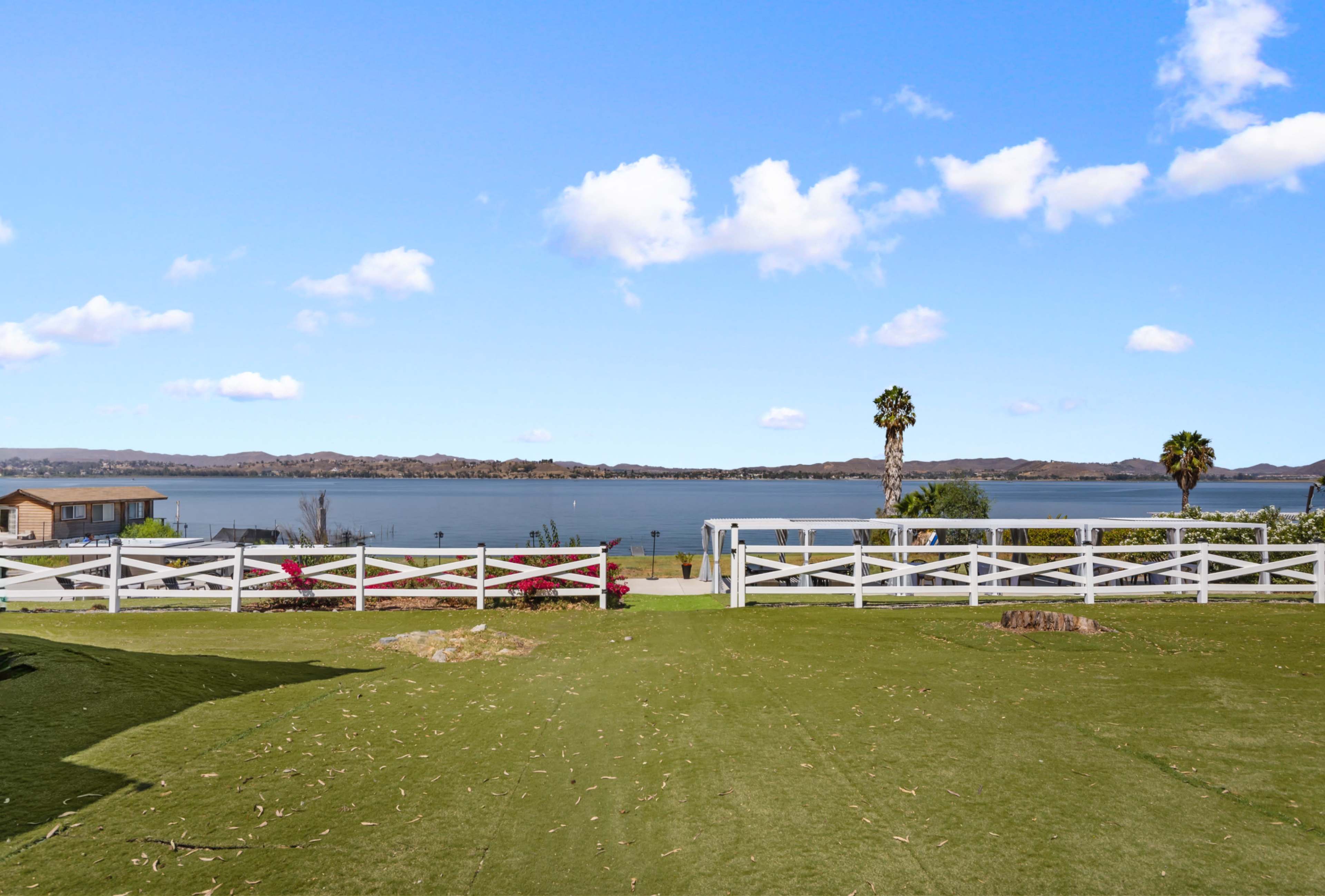 The image shows a green lawn area leading to a calm body of water with hills in the background and a few structures beside the shore.
