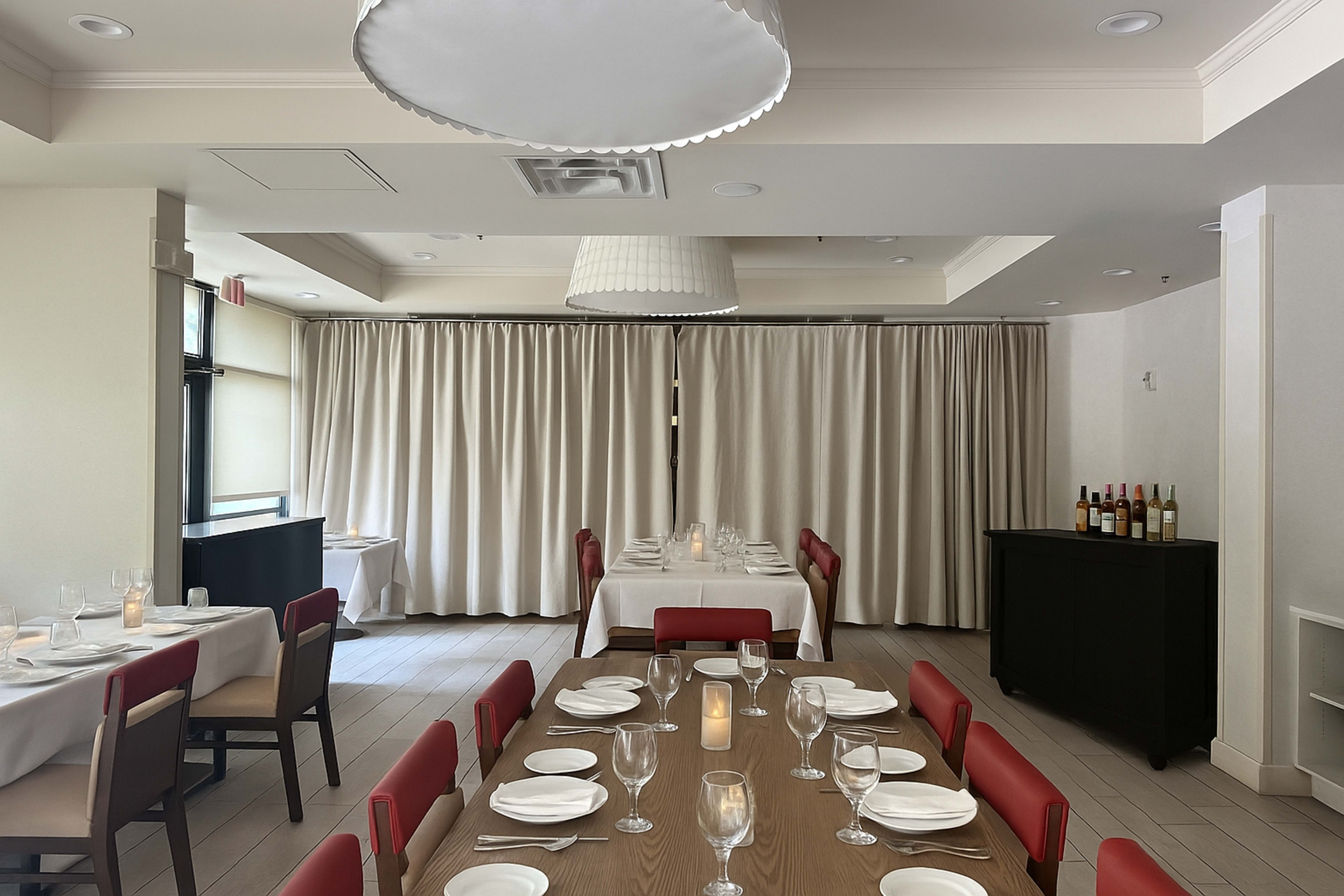 The image shows a well-organized dining area in a restaurant, featuring tables set with white tablecloths and red chairs, alongside a wine rack against the wall.