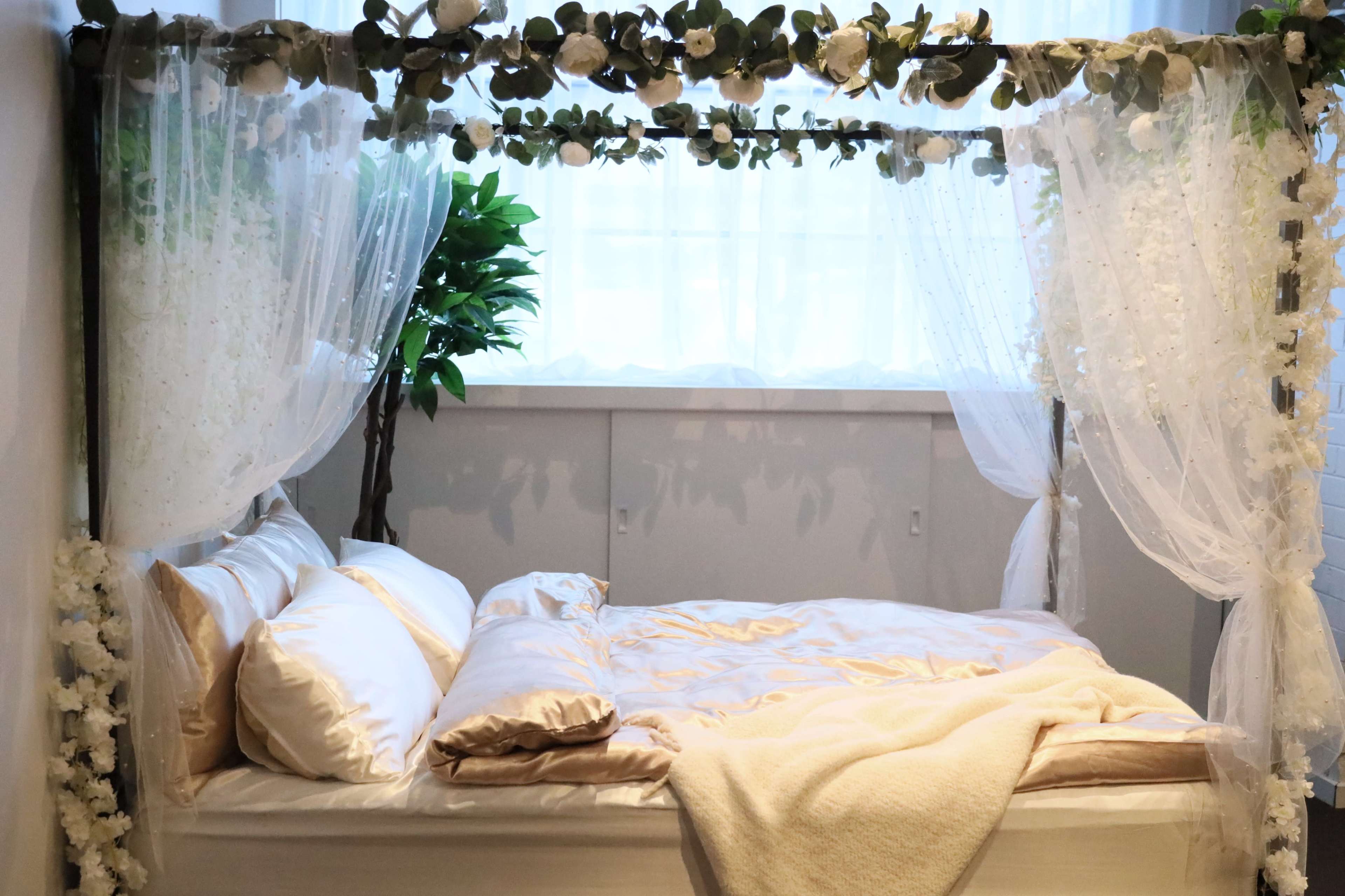 A four-poster bed with sheer curtains and decorative greenery is positioned near a window.