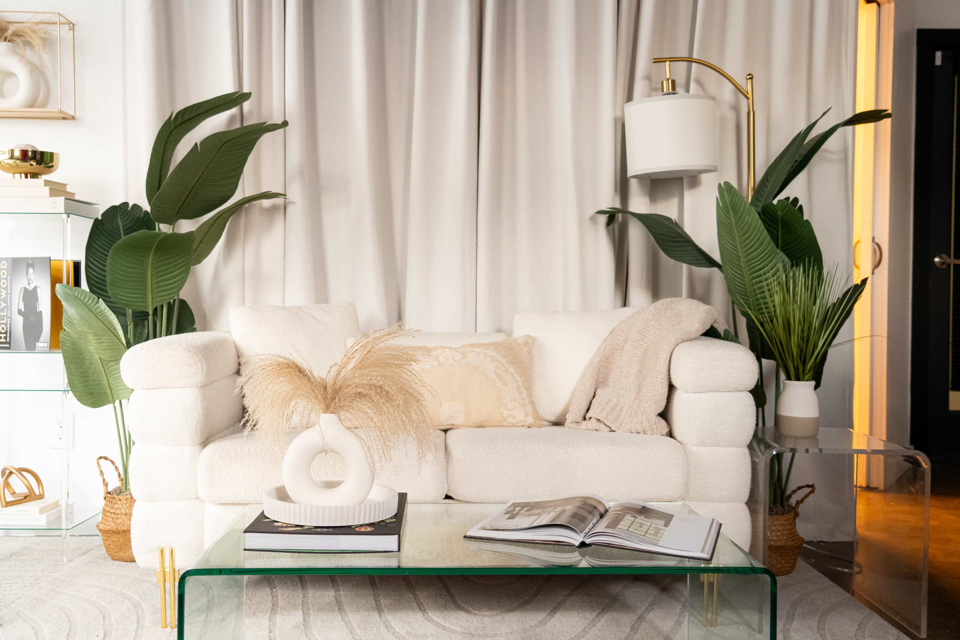 A cozy living room features a white sofa adorned with pillows, a glass coffee table with a magazine, and large green plants on either side.