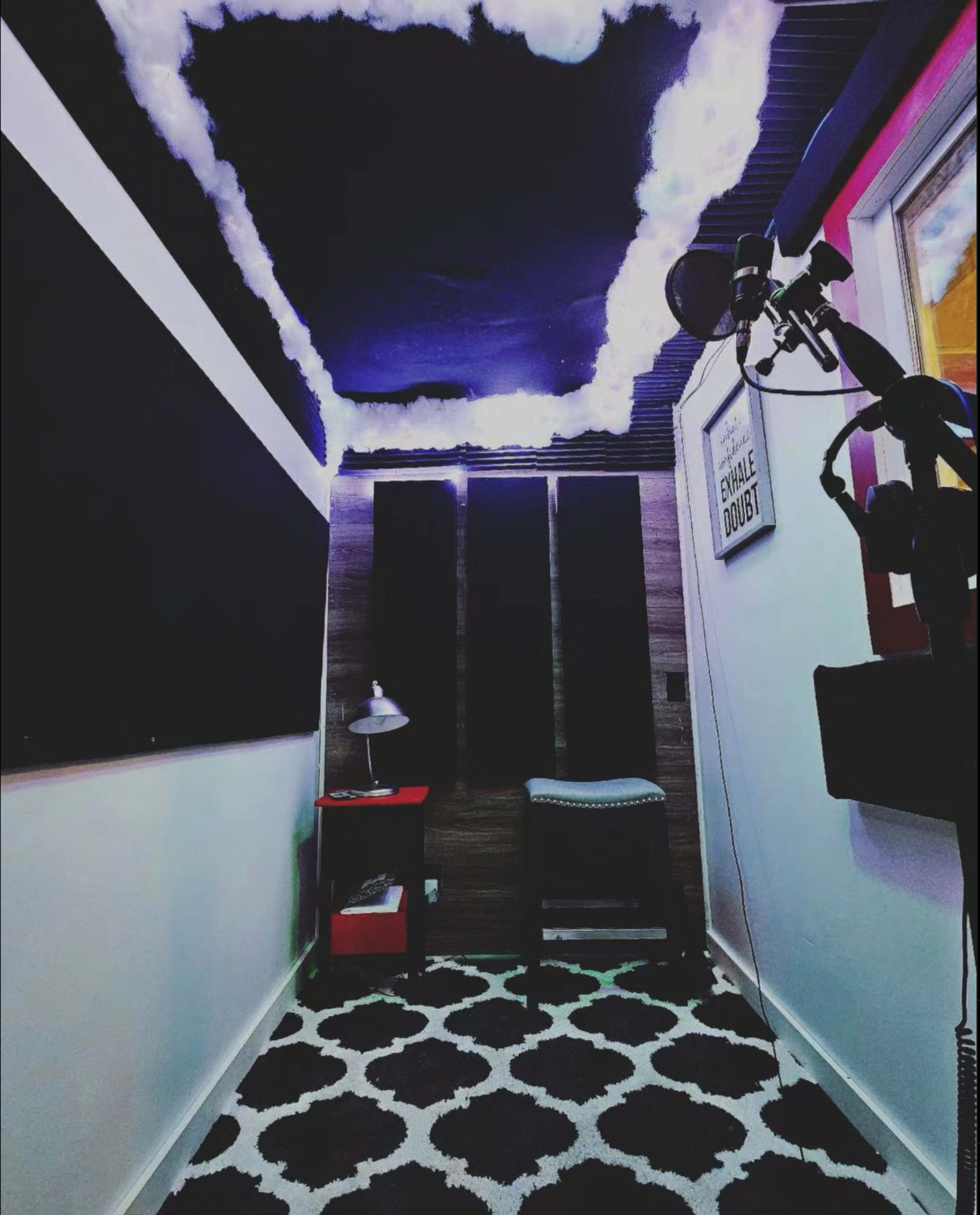 A small room designed for recording, featuring a dark cloud-themed ceiling, a black and white rug, a lamp on a side table, and soundproofing panels on the walls.