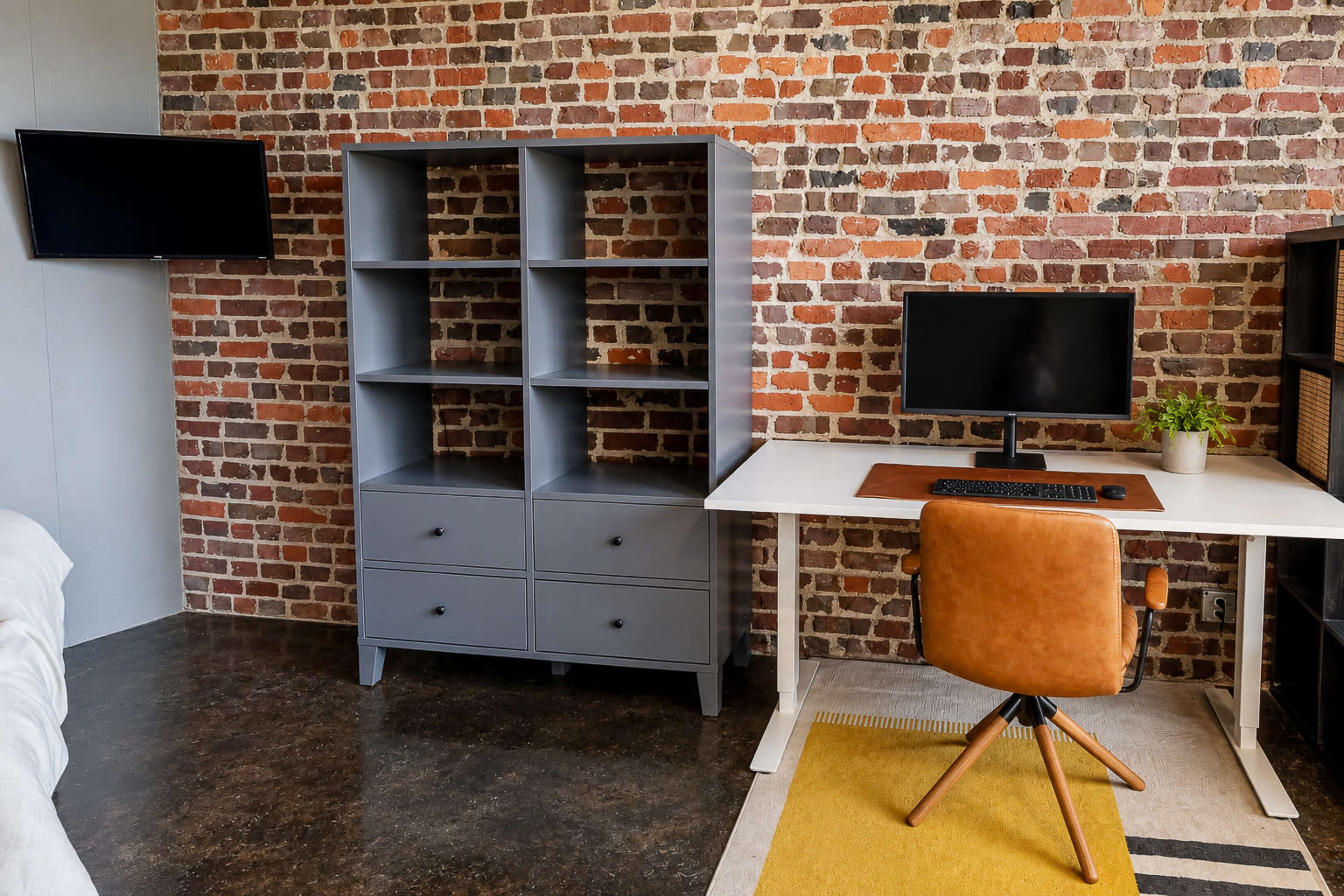 The scene shows a minimalist workspace featuring a white desk and an ergonomic chair, a gray bookshelf with drawers, a monitor, and a brick wall background.