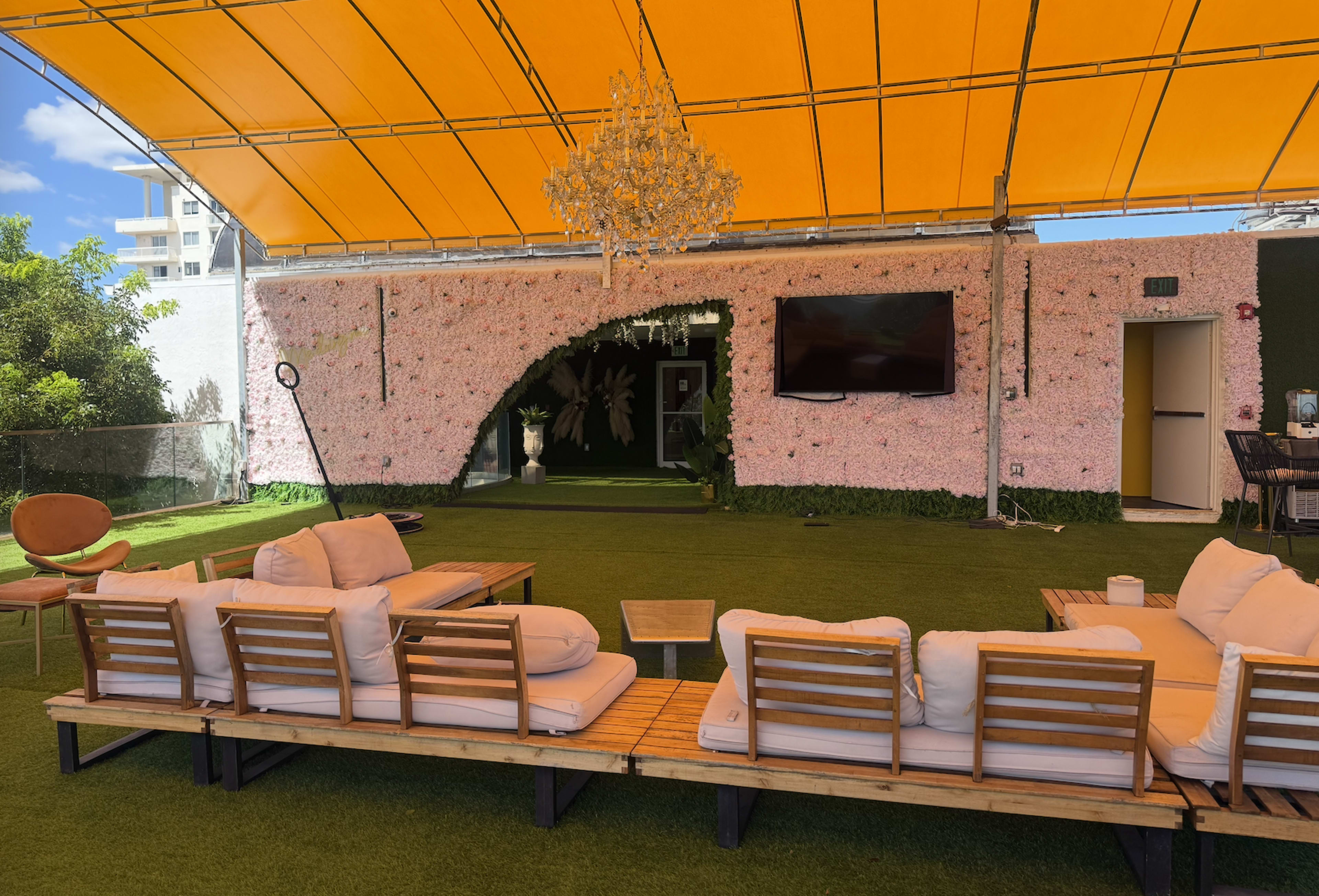 An outdoor lounge area features a large sofa with wooden frames, a glass table, and a pink flower-covered wall under a bright orange canopy.