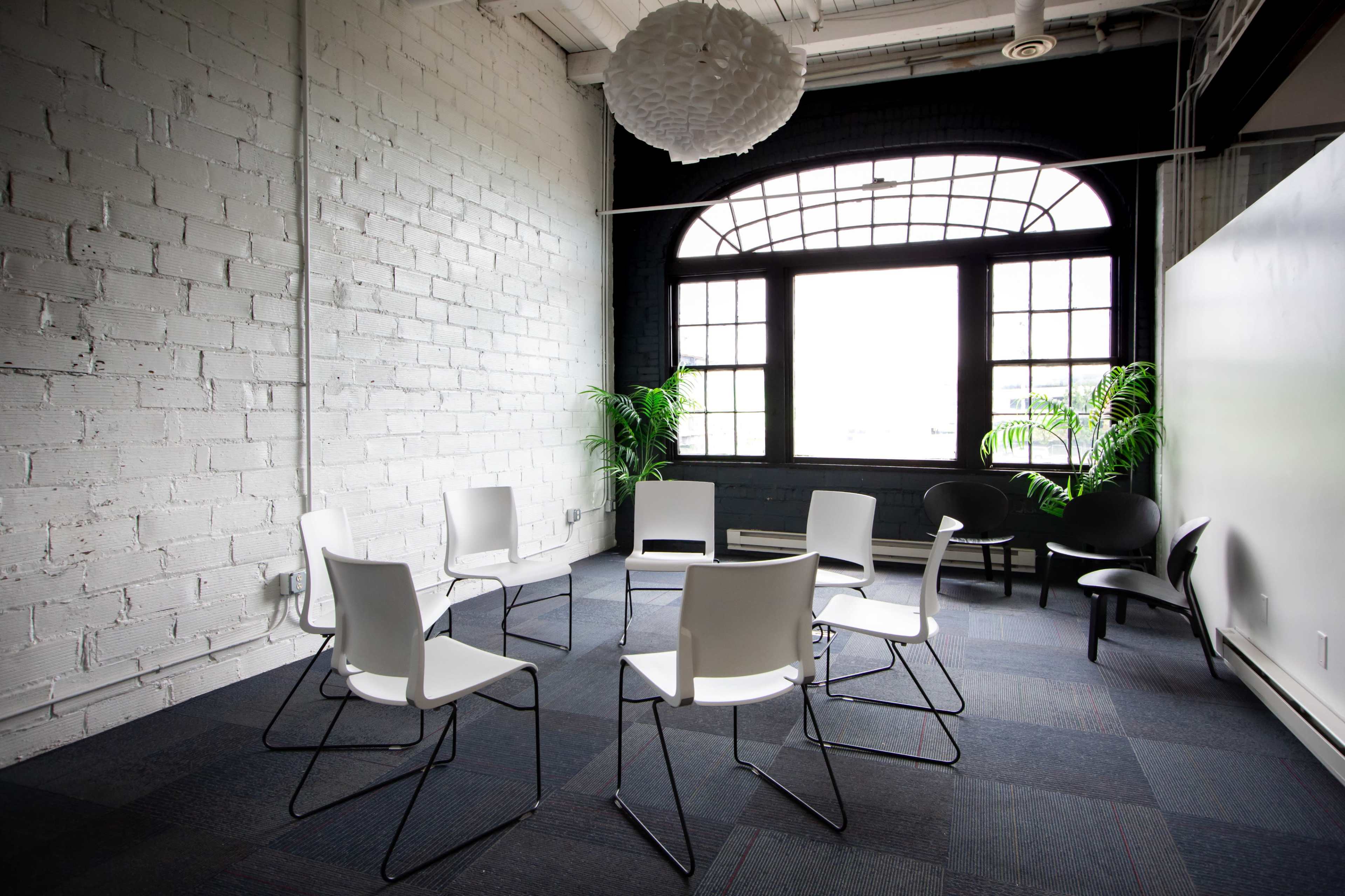 A modern, minimalist meeting room features a circular arrangement of white and black chairs around a central space, with large windows allowing natural light to enter.