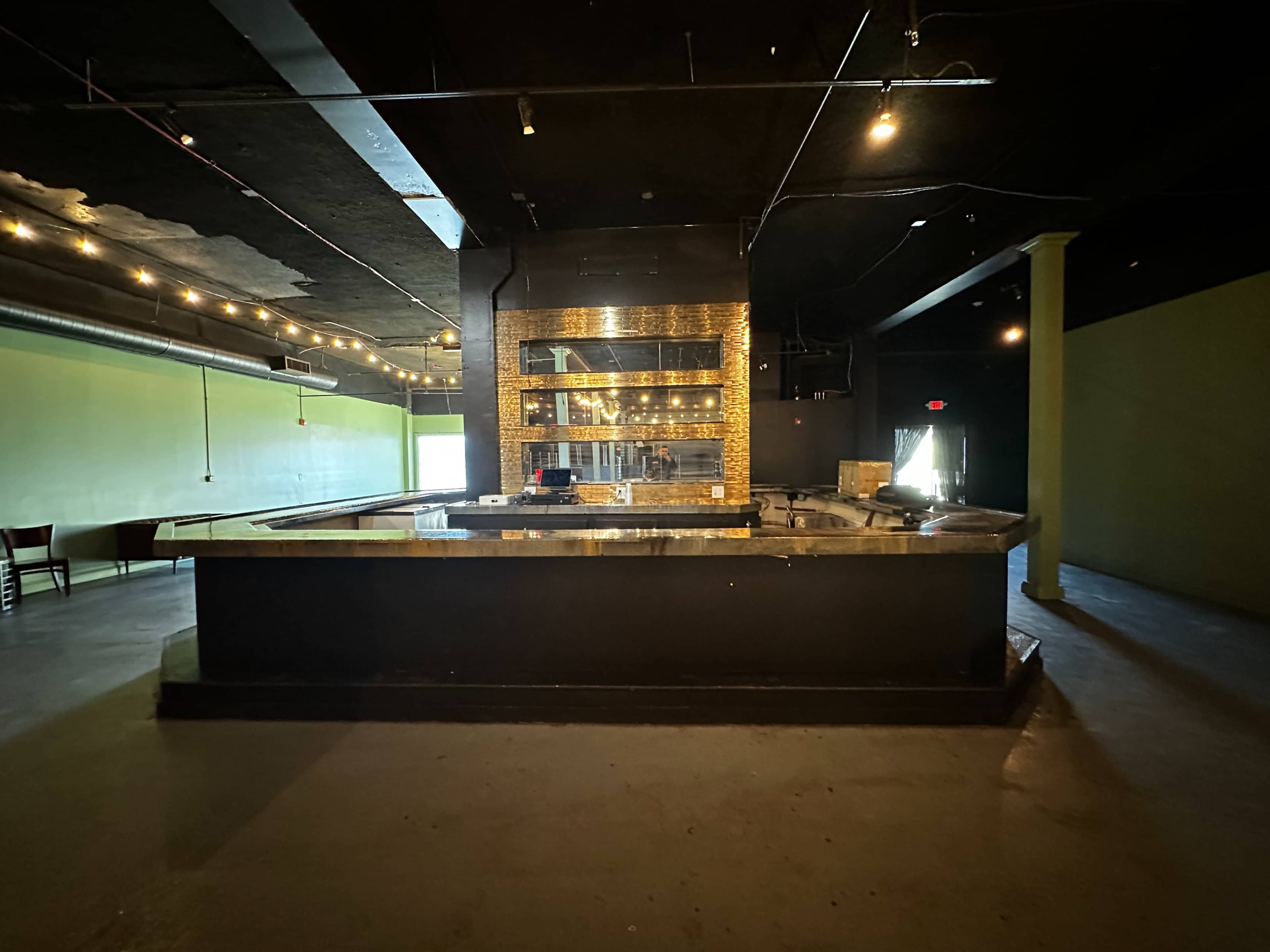 An empty bar area with a countertop and a back wall featuring shelves, set in a darkly lit room with green walls.