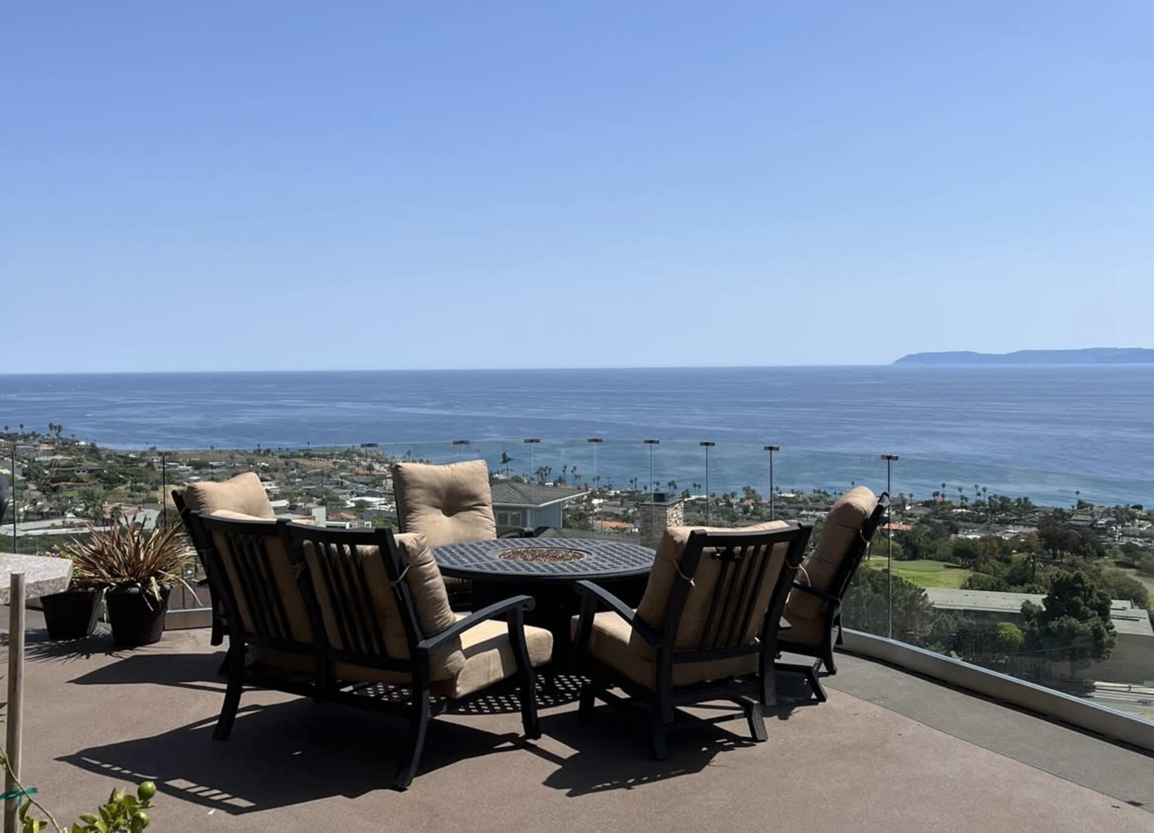 A patio table with four chairs overlooks the ocean and a coastal area under a clear blue sky.