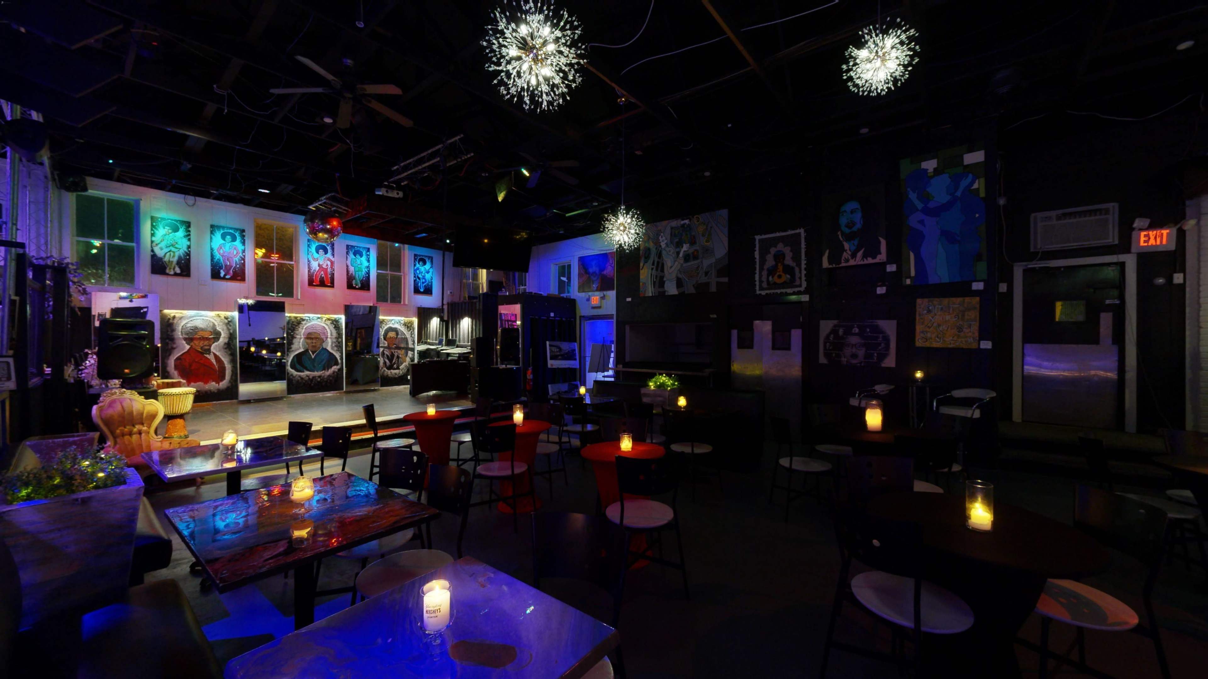 A dimly lit lounge featuring colorful artwork on the walls, tables with candles, and a small stage at the front.
