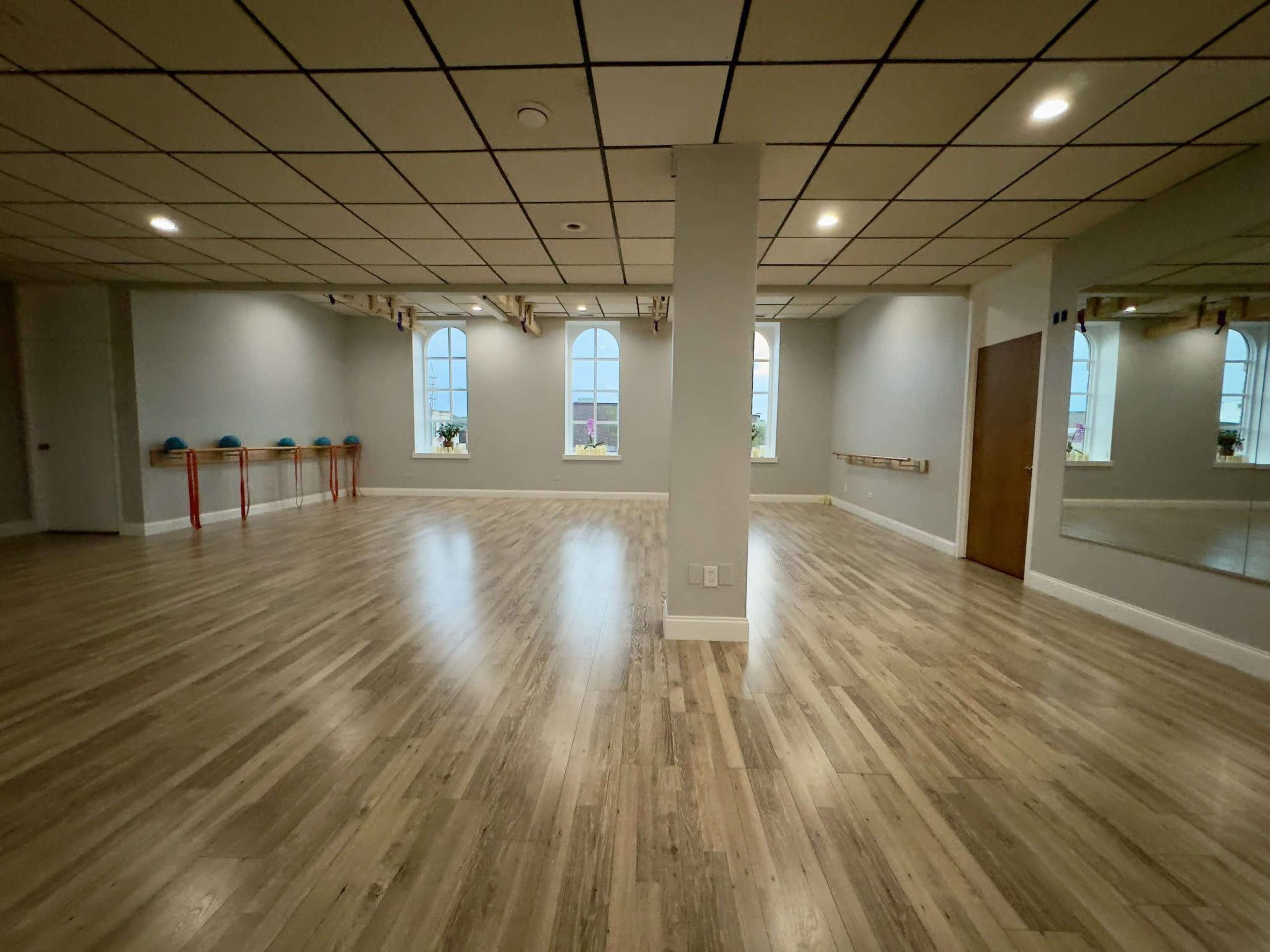 A spacious, well-lit studio with wooden flooring, mirrored walls, and large windows, featuring exercise equipment along one side.