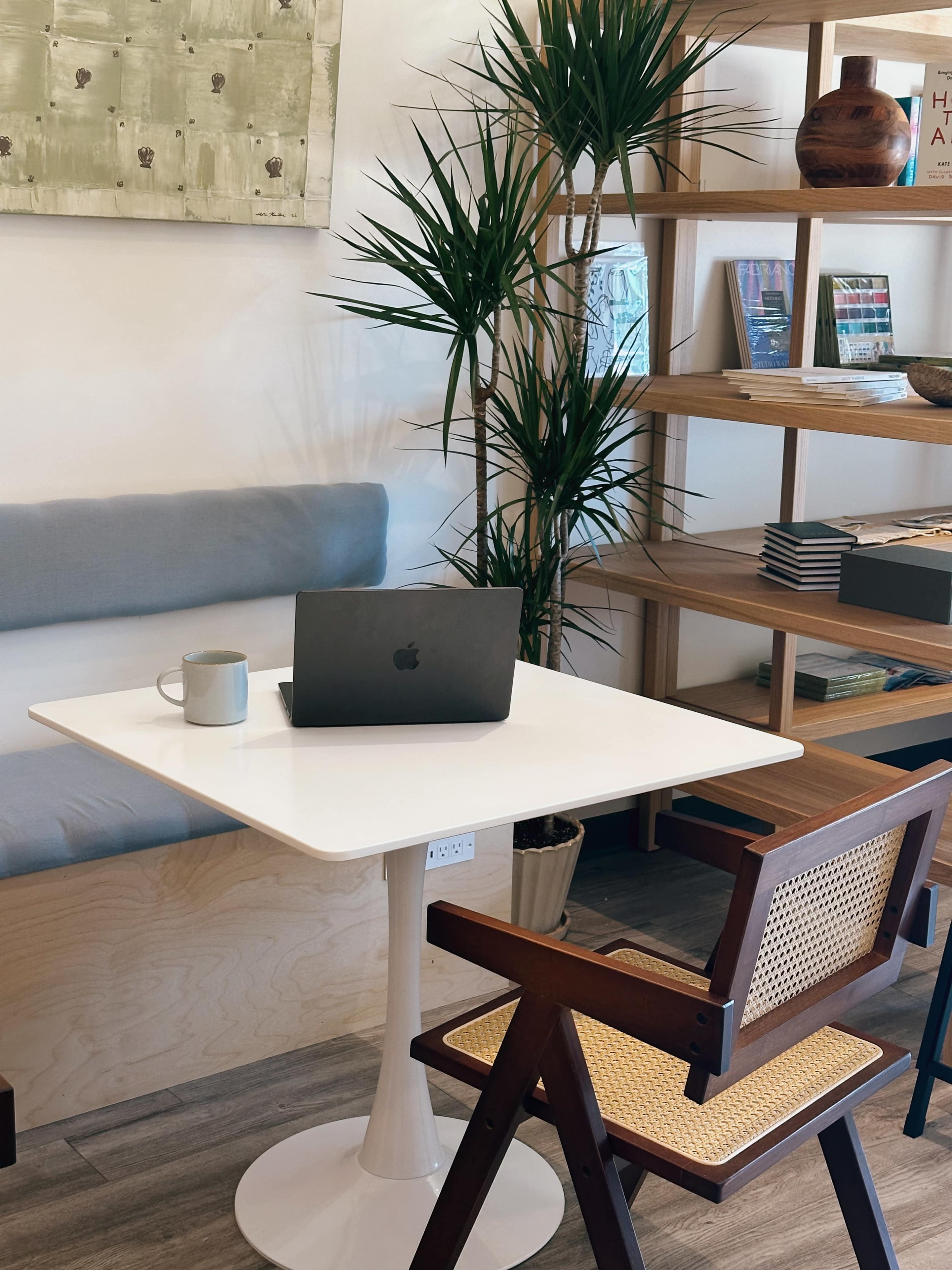 A minimalist workspace features a white table with a laptop, a mug, and a wooden chair, surrounded by potted plants and shelves filled with books and decorative items.