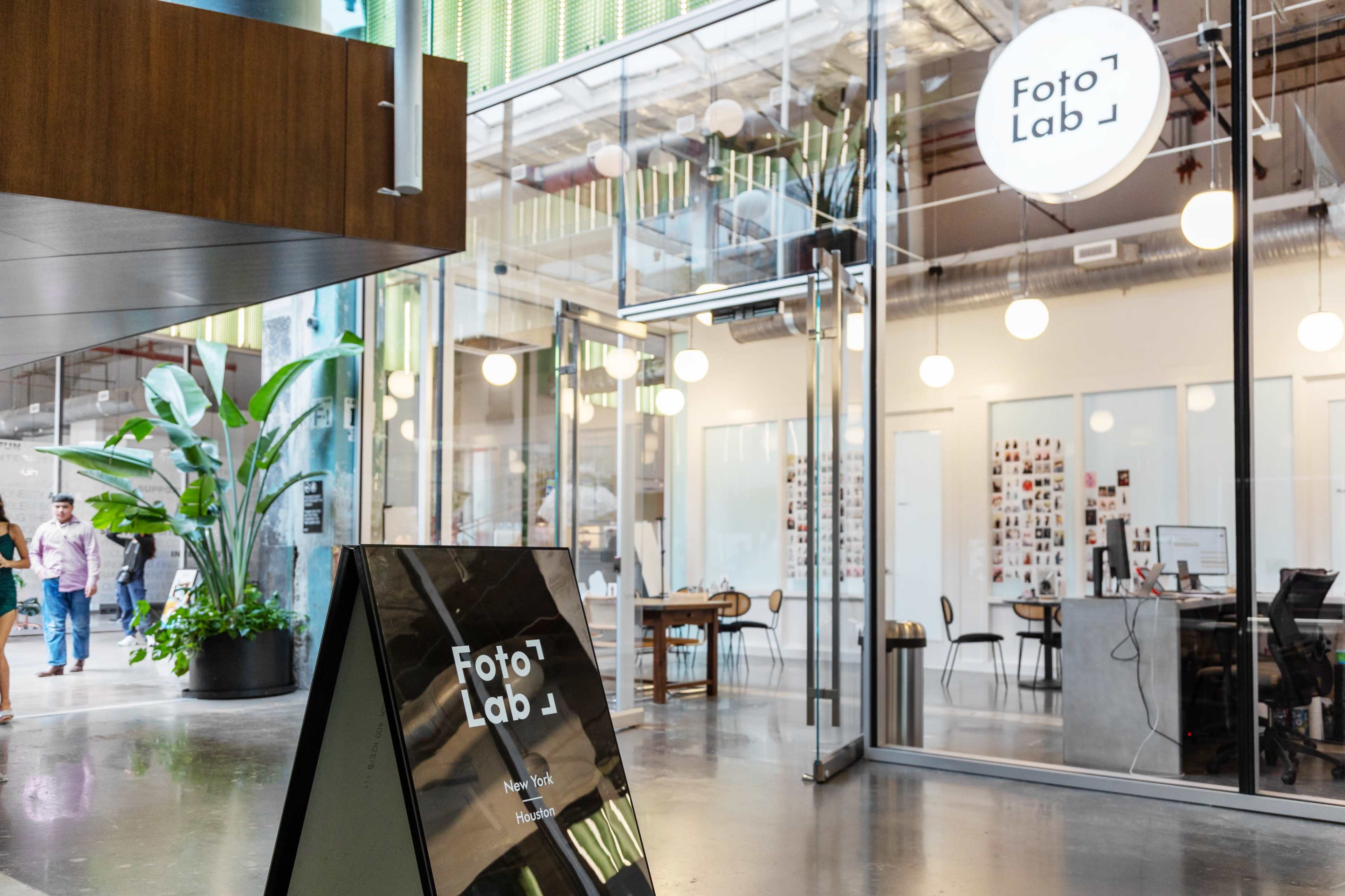 A modern interior of a space labeled "Foto Lab," featuring a large glass entrance, greenery, and a workspace with various photographs on the walls.