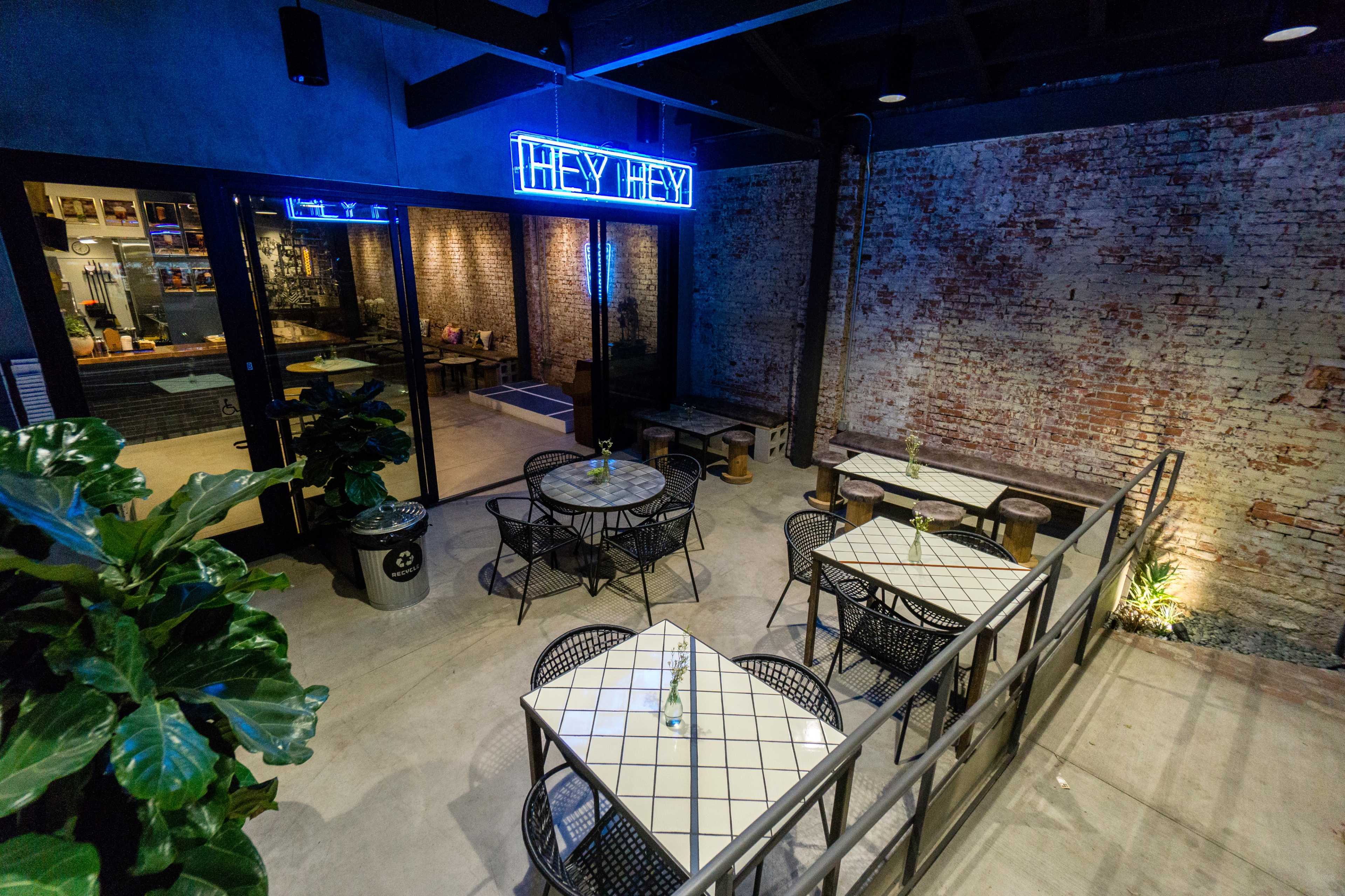 An outdoor dining area with metal tables and chairs, a brick wall, and a neon sign that says "HEY HEY."
