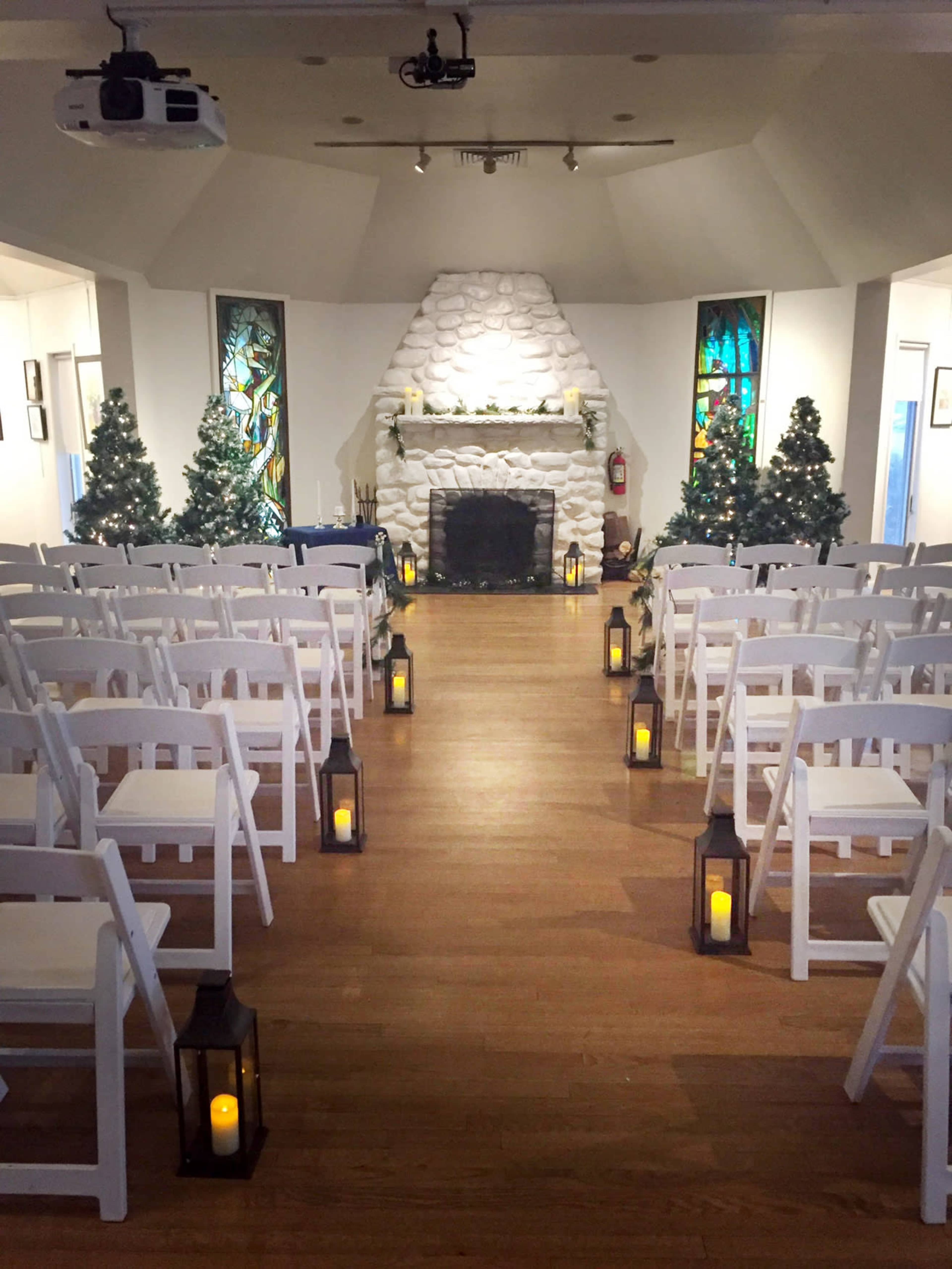 The image shows a wedding ceremony setup in a venue, with rows of white chairs facing a stone fireplace adorned with decorative elements, flanked by Christmas trees and candle lanterns along the aisle.