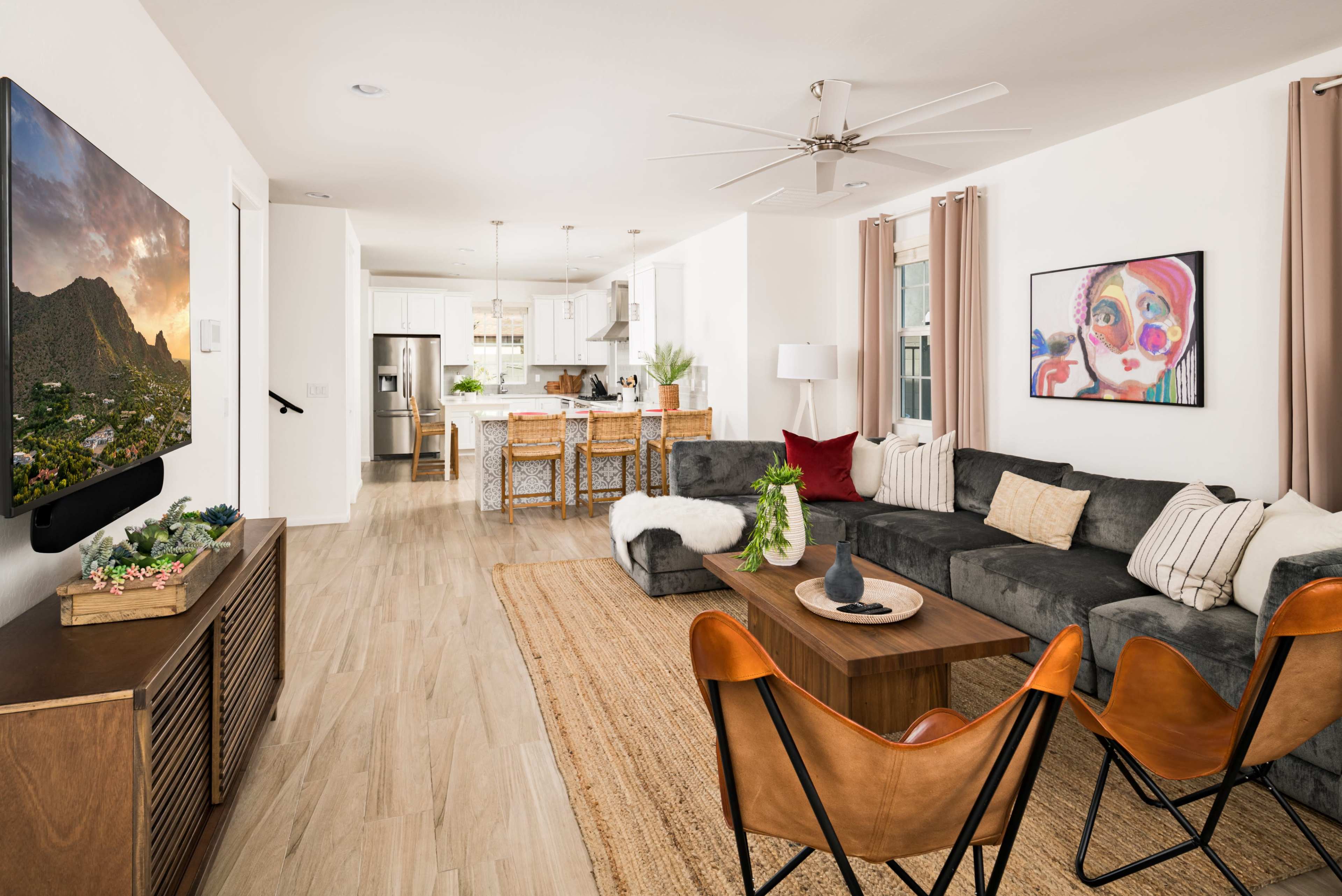 The living room features a dark gray sectional sofa, a wooden coffee table, and a wall-mounted television, with a kitchen area visible in the background.