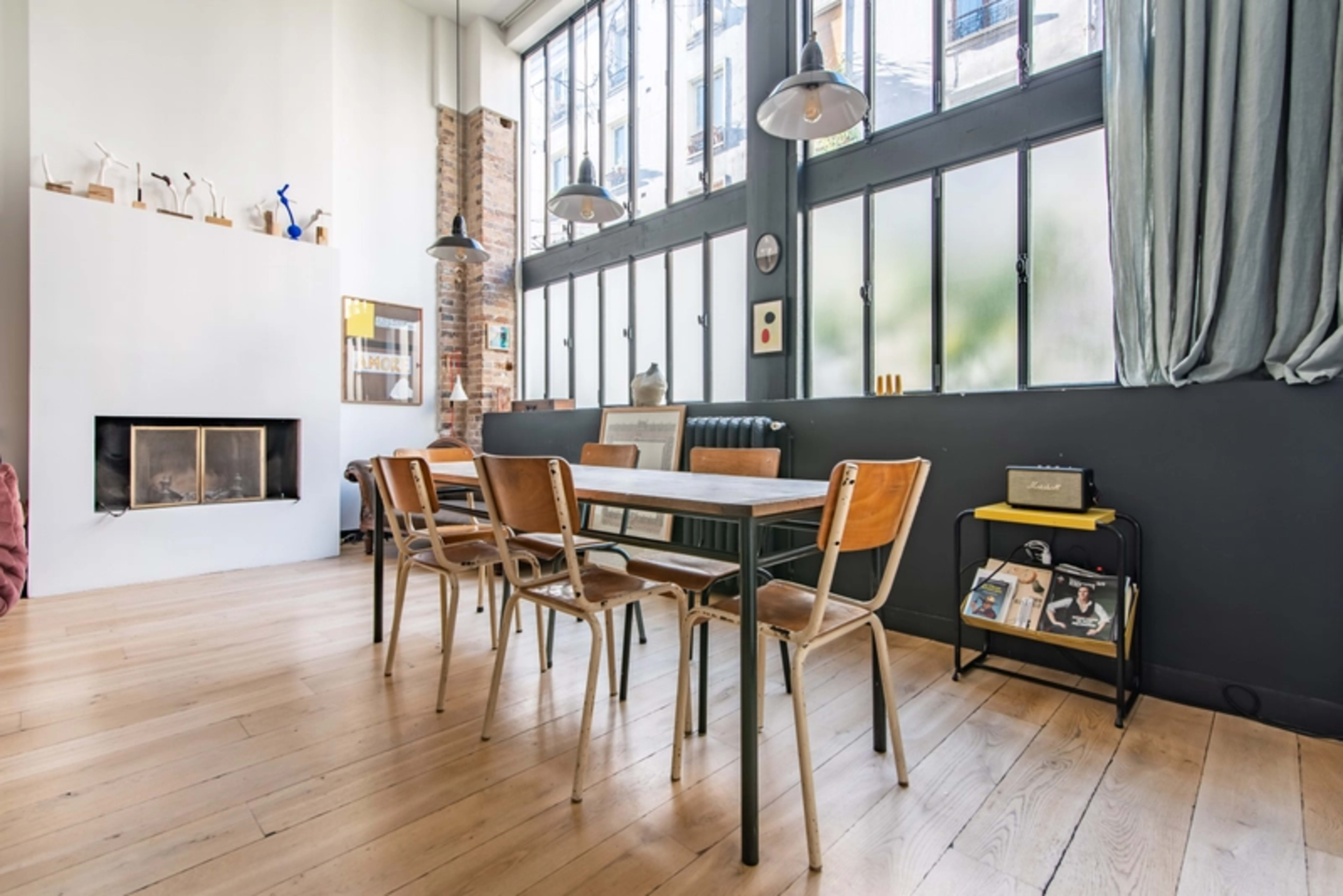 A dining area features a wooden table surrounded by chairs, large windows allowing natural light, and a fireplace against one wall.