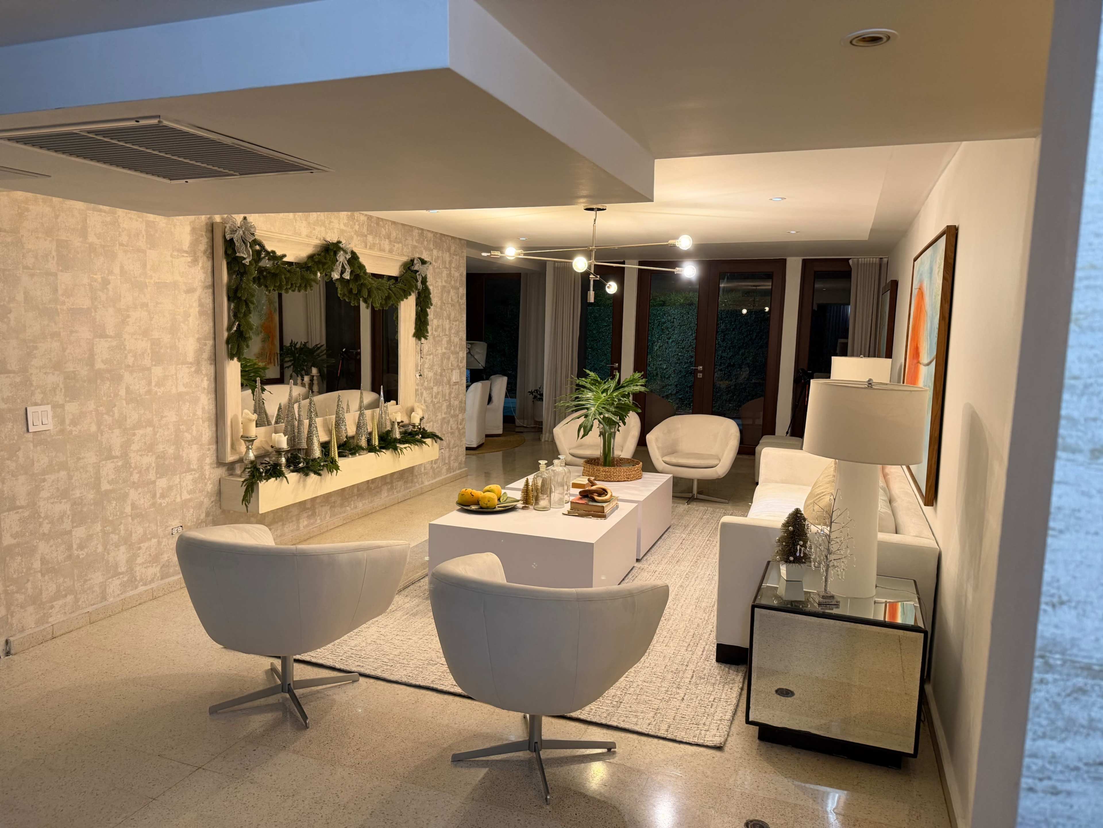 The image shows a modern living room with white furniture, a coffee table, decorative greenery, and large windows displaying an outdoor view.