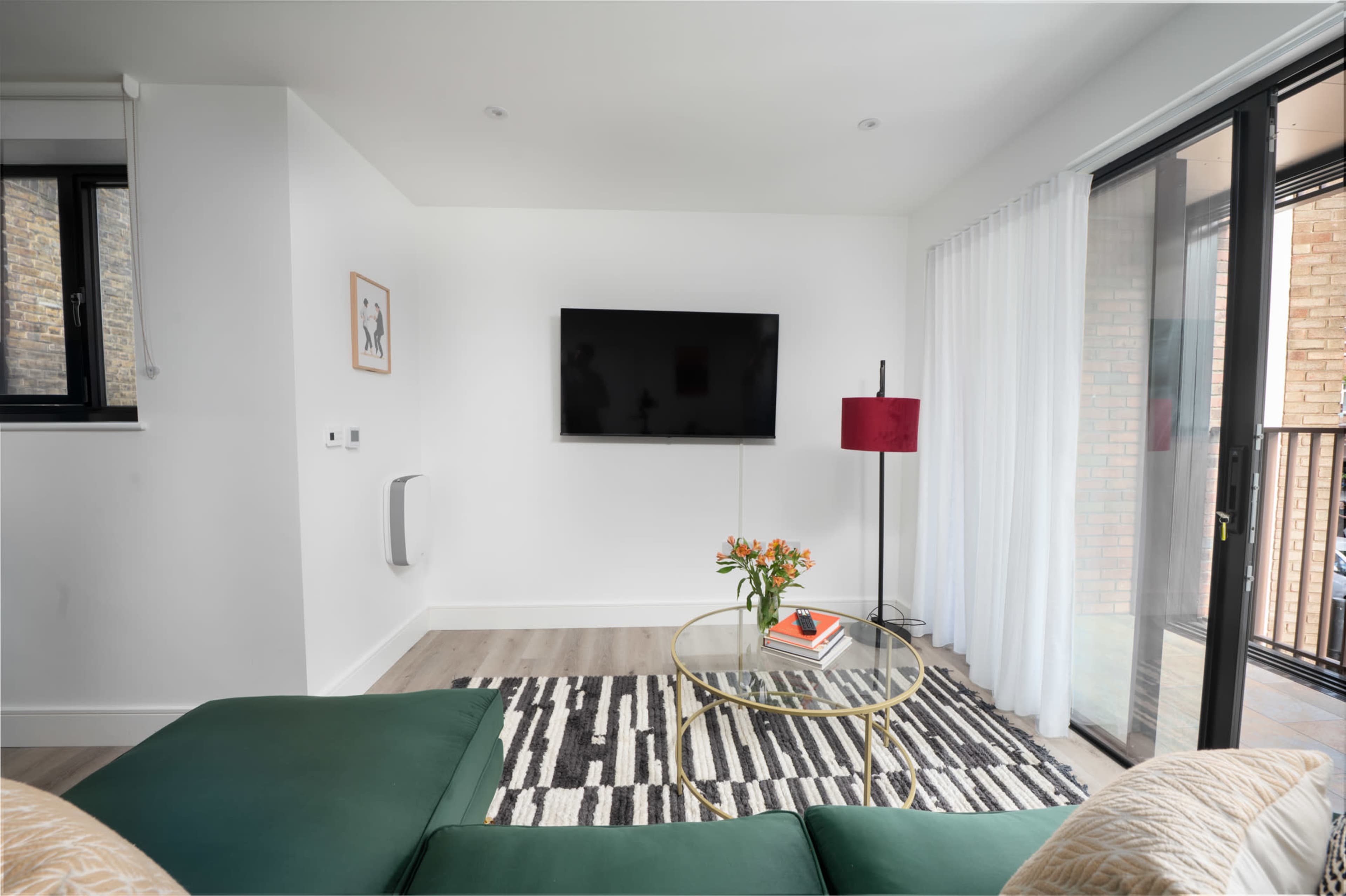 The scene features a modern living room with a green sectional sofa, a round glass coffee table, a television mounted on the wall, and a floor lamp beside a window with sheer curtains.