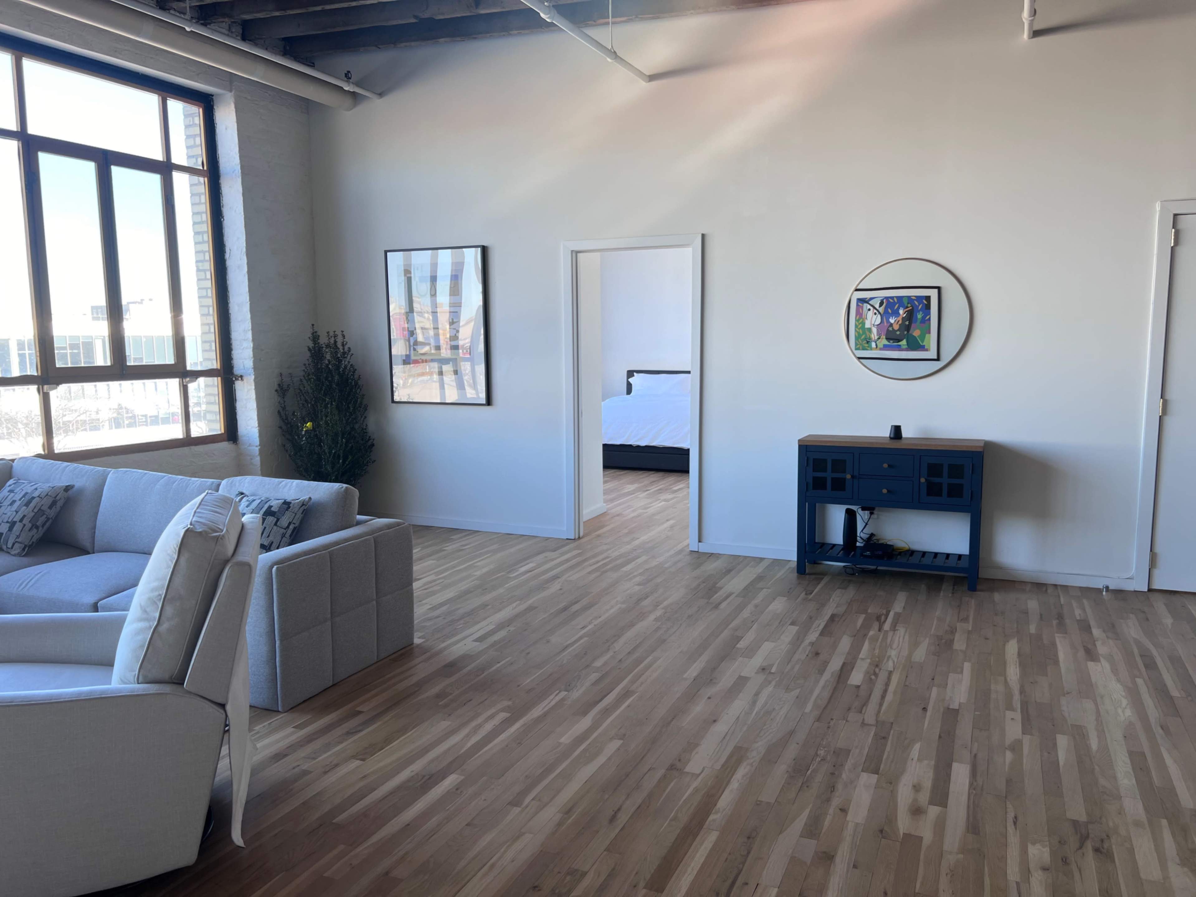 The image shows a spacious, modern room with a gray sofa, a blue console table, and an open doorway leading to a bedroom.