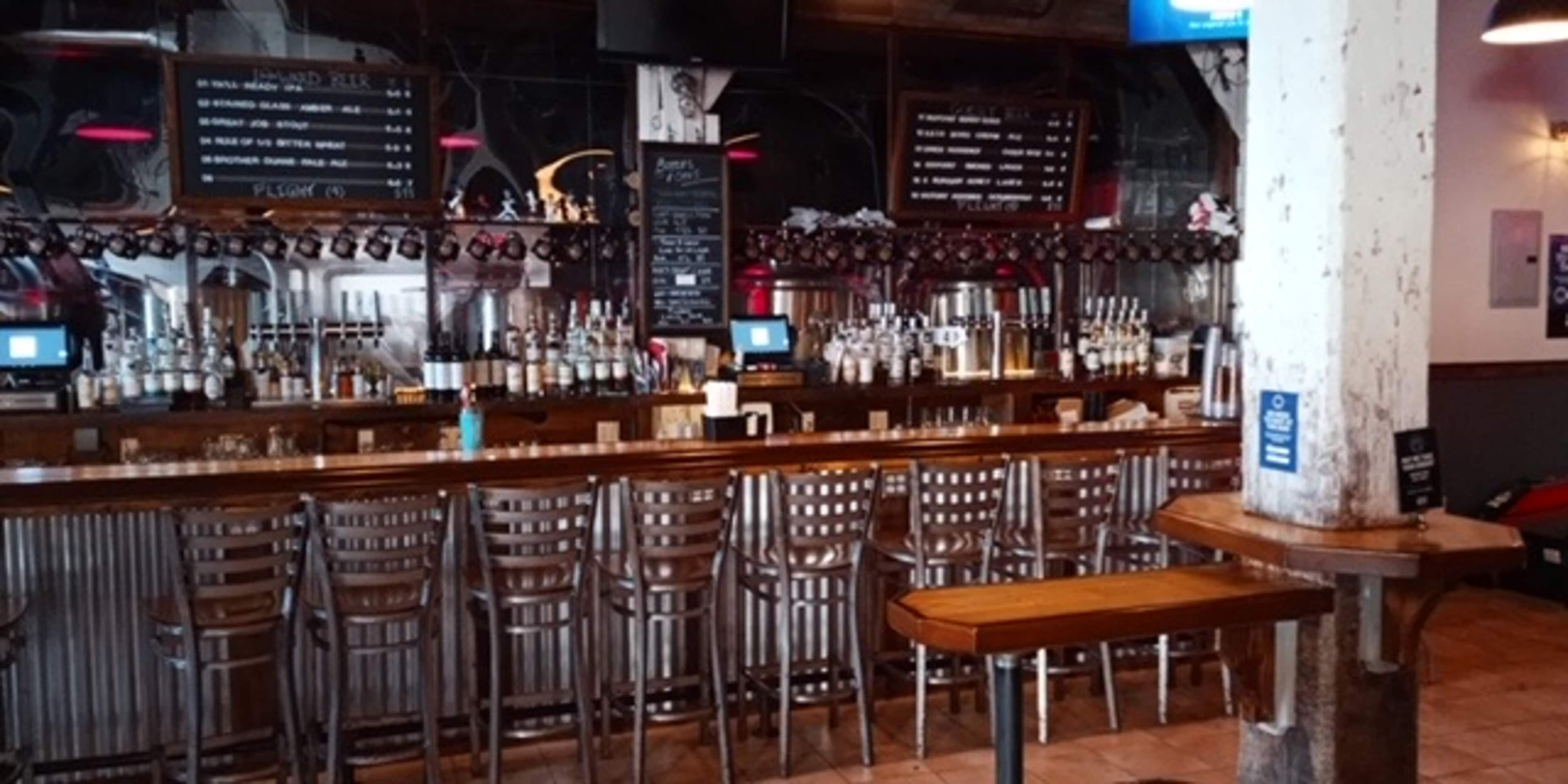 The interior of a bar with a long wooden counter, high stools, and shelves lined with various bottles and taps in the background.