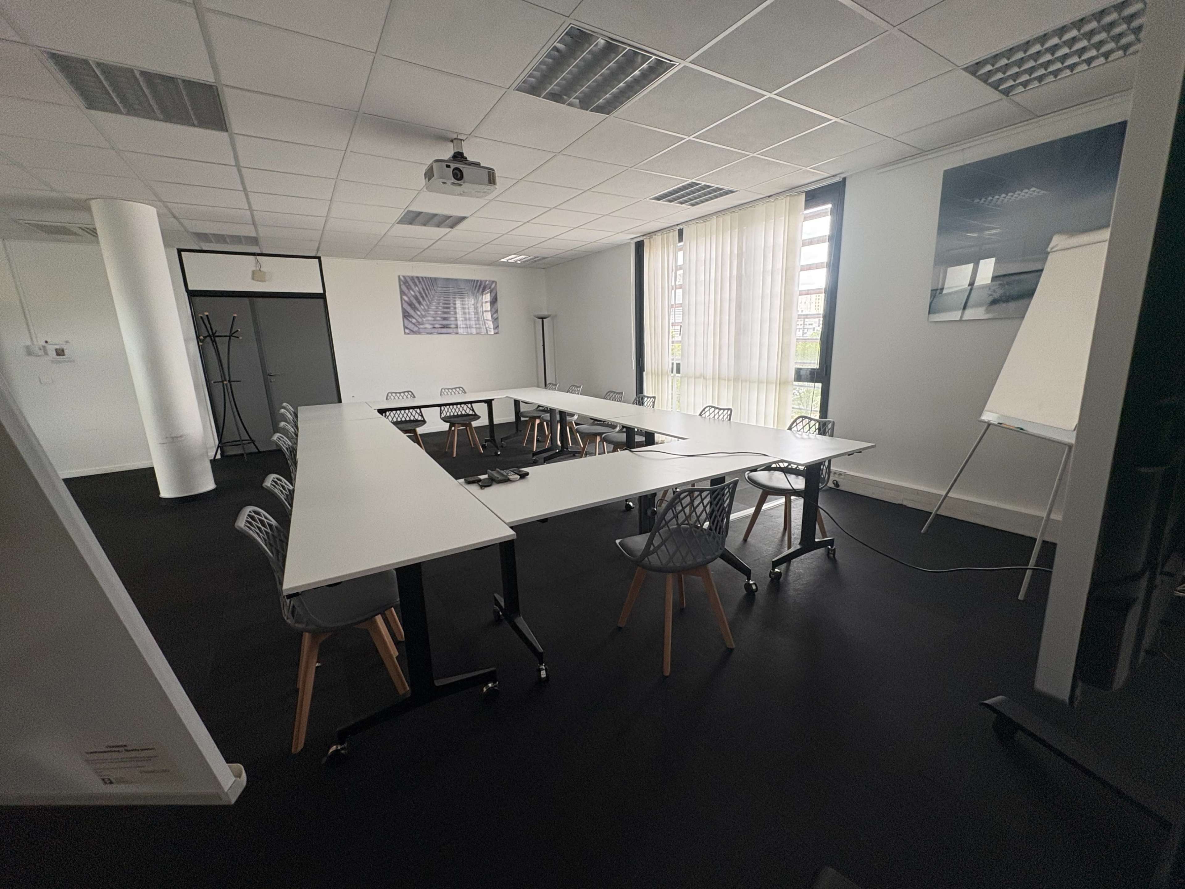 The image shows a modern conference room set up with white tables arranged in a U-shape, surrounded by chairs, with large windows letting in natural light.